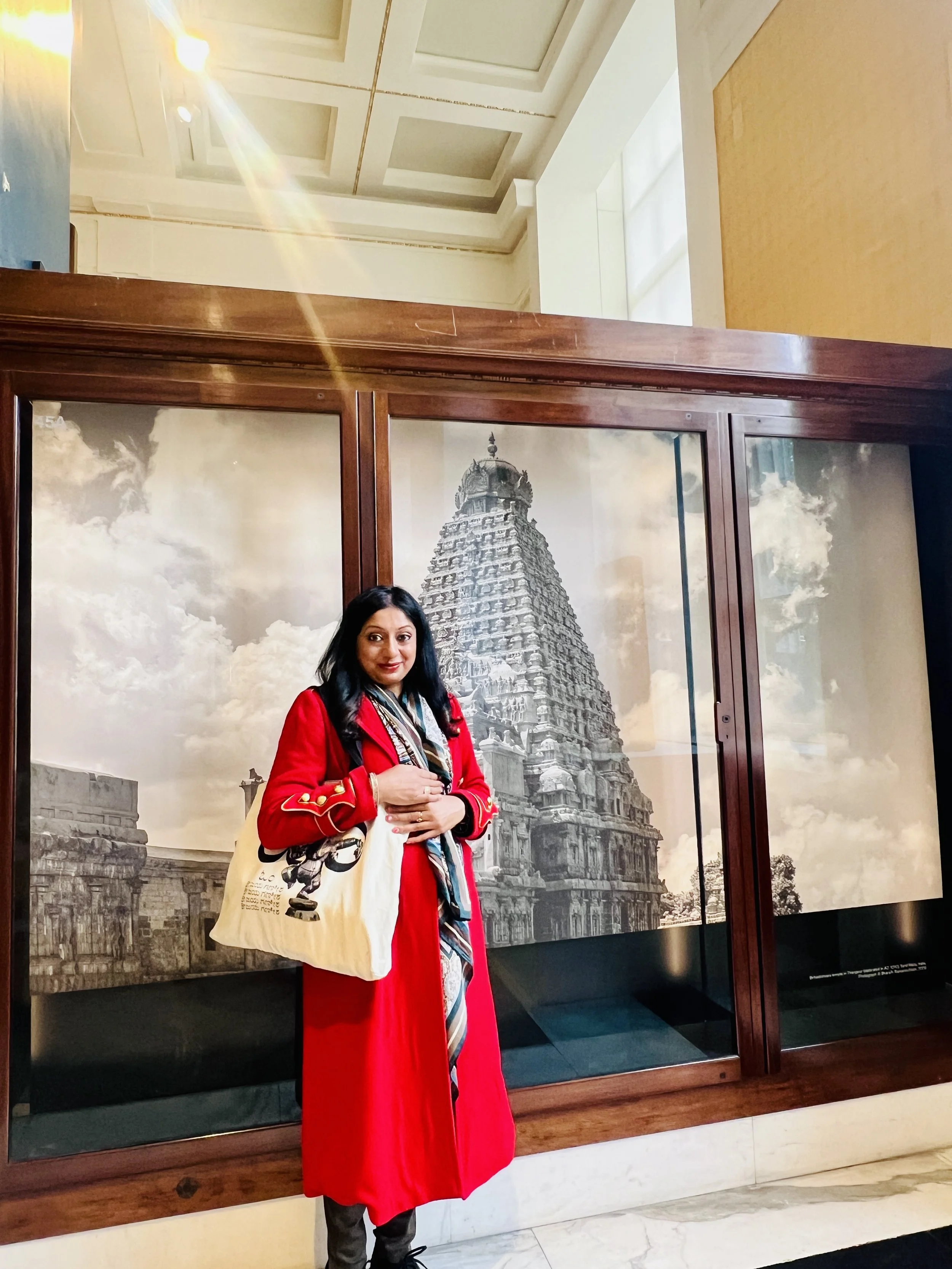 Designer and Hindu Chaplain Sangeetha Kowsik in front of an image of traditional South Indian Hindu temple architecture, at the British Museum, carrying one of her Ihsan Ishan Design art bags.