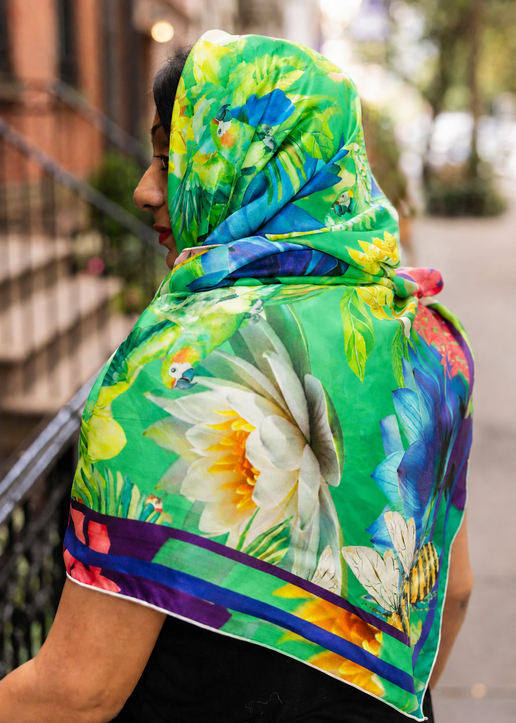 Model wearing a green botanical silk scarf by Ihsan Ishan Design featuring parrots, tropical flowers and bees, styled as a headscarf and wrap.