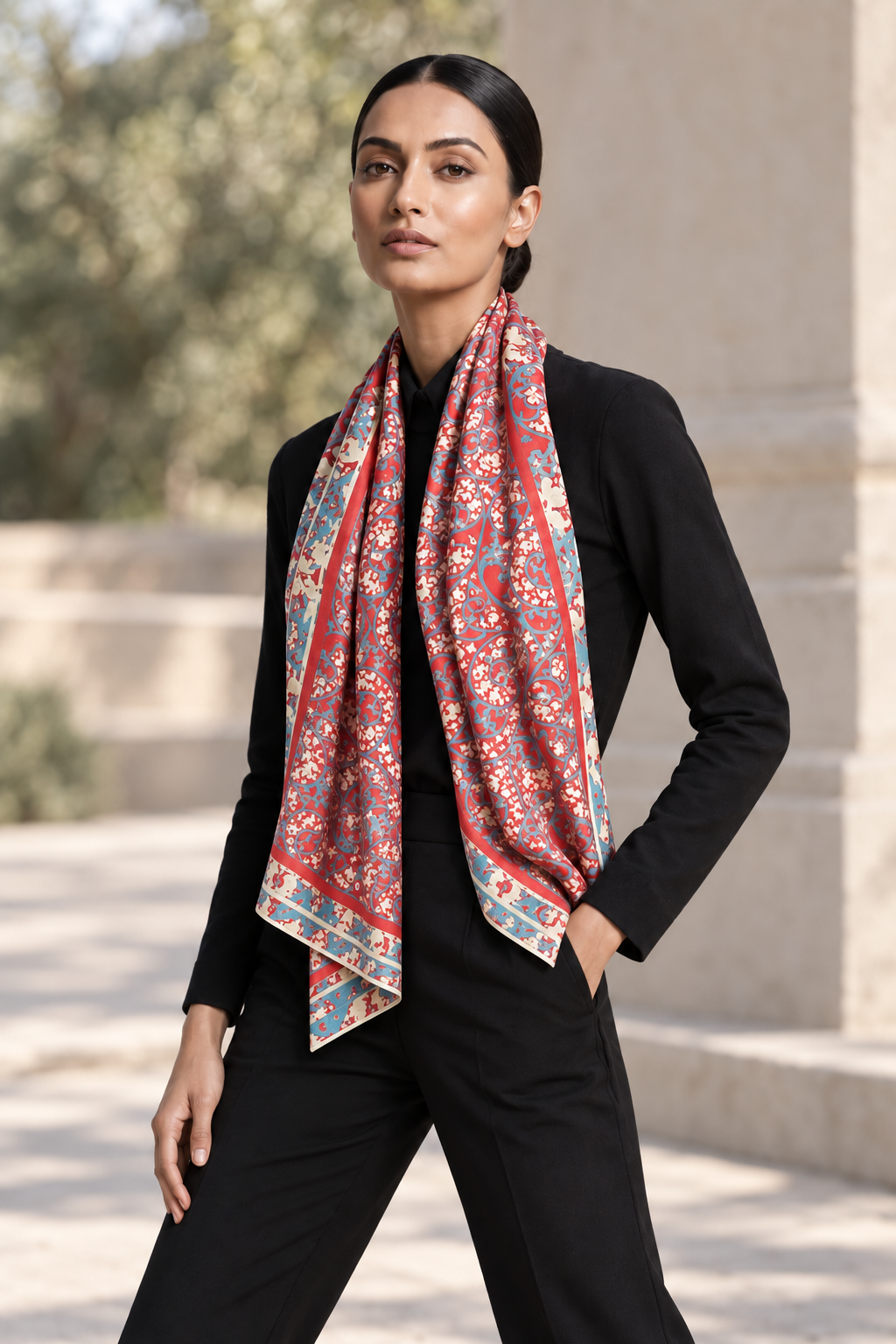 Model in a black outfit wearing a red Persian silk scarf with turquoise and ivory floral motifs, casually draped around her neck outdoors.