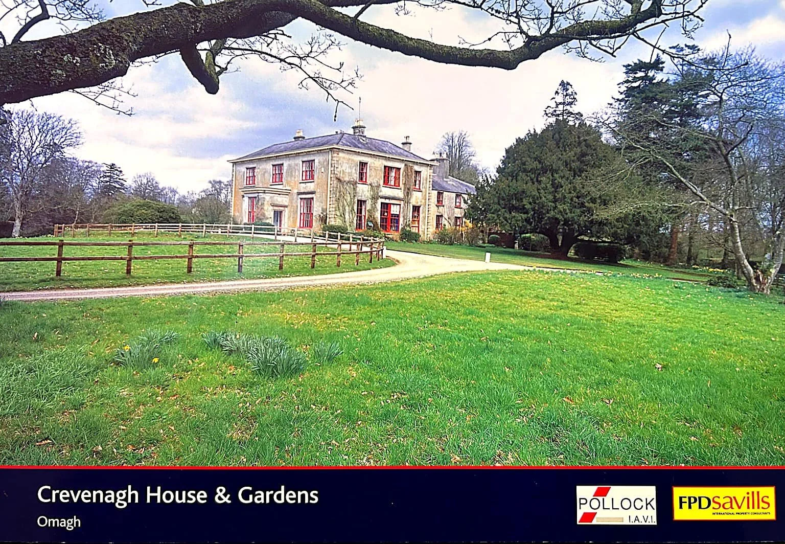 A large historic stone house with multiple red windows is situated on a lush green lawn with yellow flowers. A generic dirt path curves in front of the house, with a wooden fence along it. Mature trees surround the property, with some bare branches and others lush with leaves, under a partly cloudy sky.