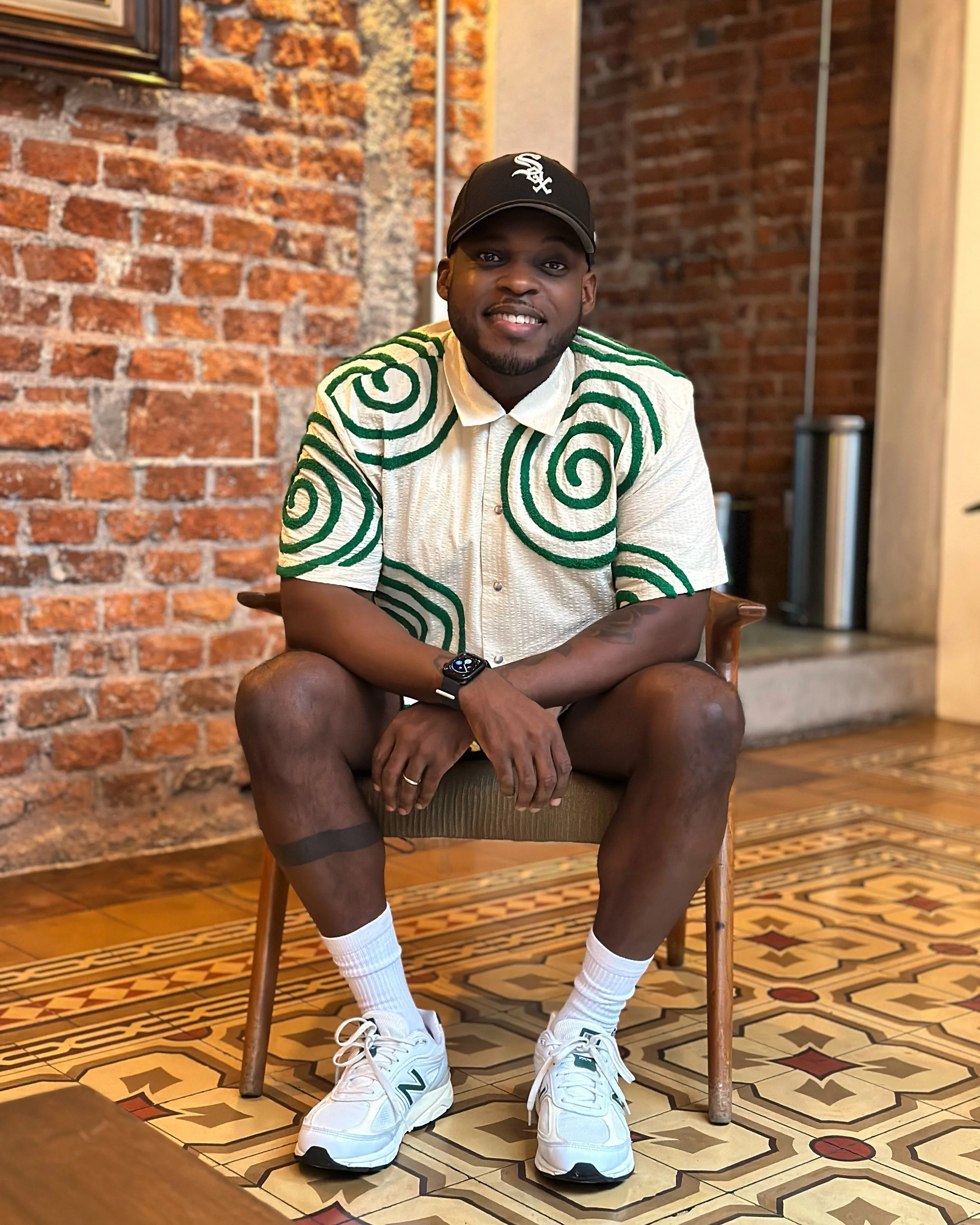 A young man sitting on a wooden chair in an indoor setting with exposed brick walls. He is wearing a black baseball cap, a cream-colored shirt with green spiral patterns on the shoulders and sleeves, white sneakers, and white socks. He is smiling and looking into the camera.
