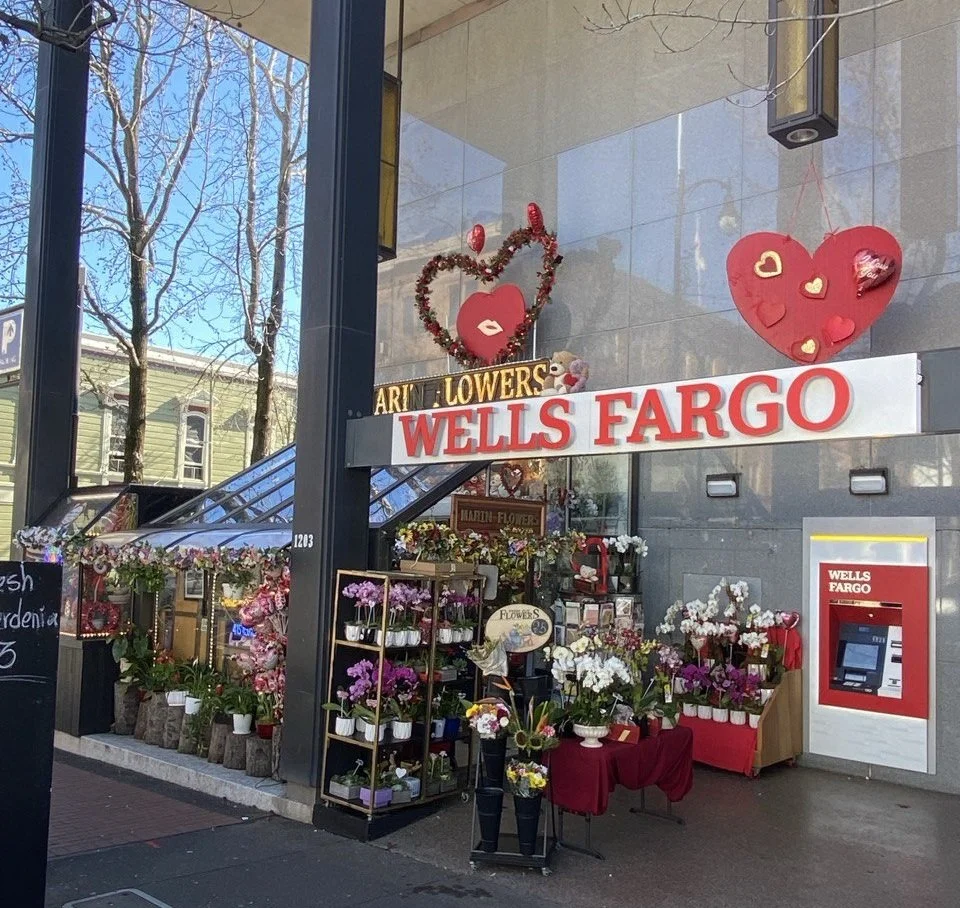 Flower shop with Valentine's Day decorations outside, displaying various flowers and red, pink, and white hearts, with a Wells Fargo ATM on the wall.
