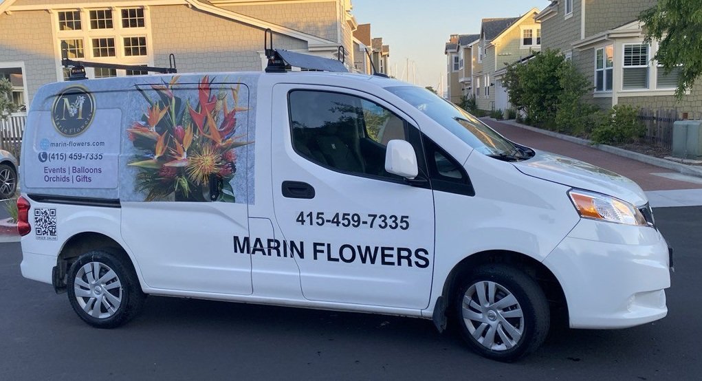White business van with Marin Flowers logo, phone number, and a floral graphic on the side, parked on a street with houses in the background