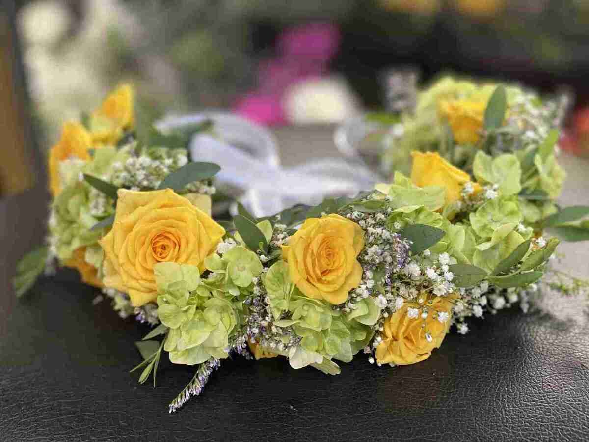 Yellow floral crown with roses, greenery, and small white flowers on a black surface.