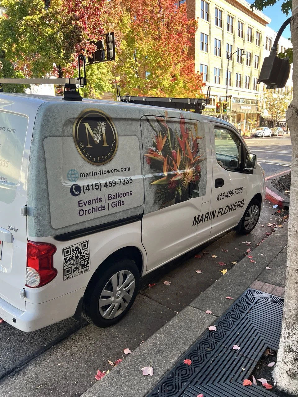 A white van parked on a city street has advertisements for Marin Flowers, featuring a logo with a black circle and gold border, a large floral image, and contact information. The surroundings include autumn leaves on the street and a building with multiple windows.