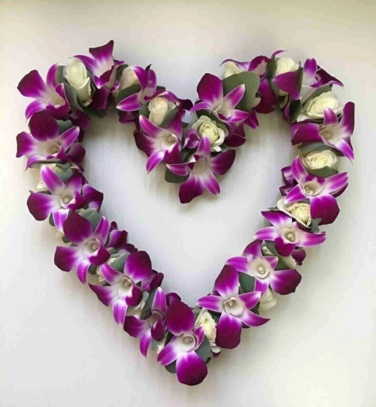 A heart-shaped arrangement of purple and white orchids with green leaves on a white background.