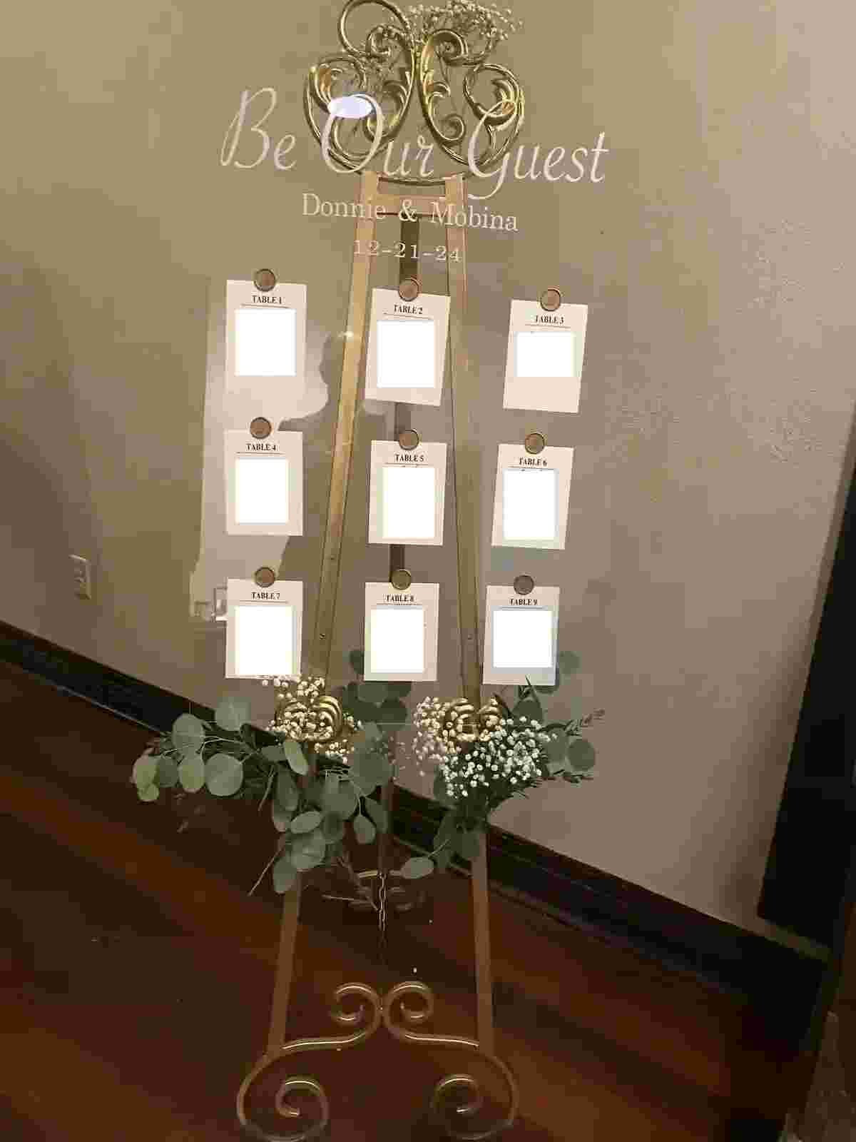 Wedding seating chart on an easel with floral decorations. The chart has spaces labeled for tables, organized in two columns, with some spaces blank or with white placeholders. The backdrop features the words 'Be Our Guest' and the names 'Donnie & Mobina' along with the date '12-21-24'.