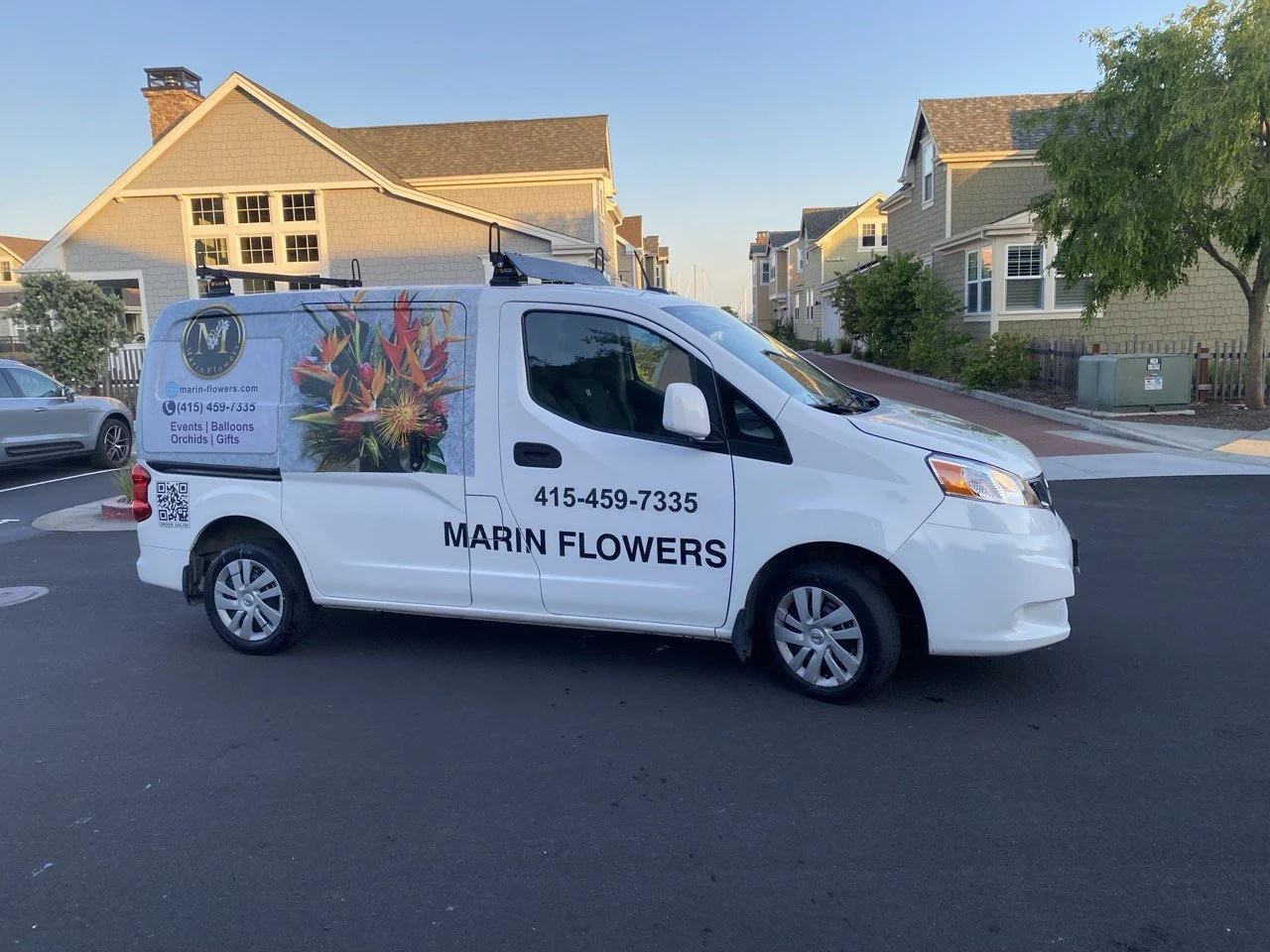 White delivery van with marigold flowers, company name, phone number, website, QR code, and services listed on side, parked on a residential street with houses and trees in the background.