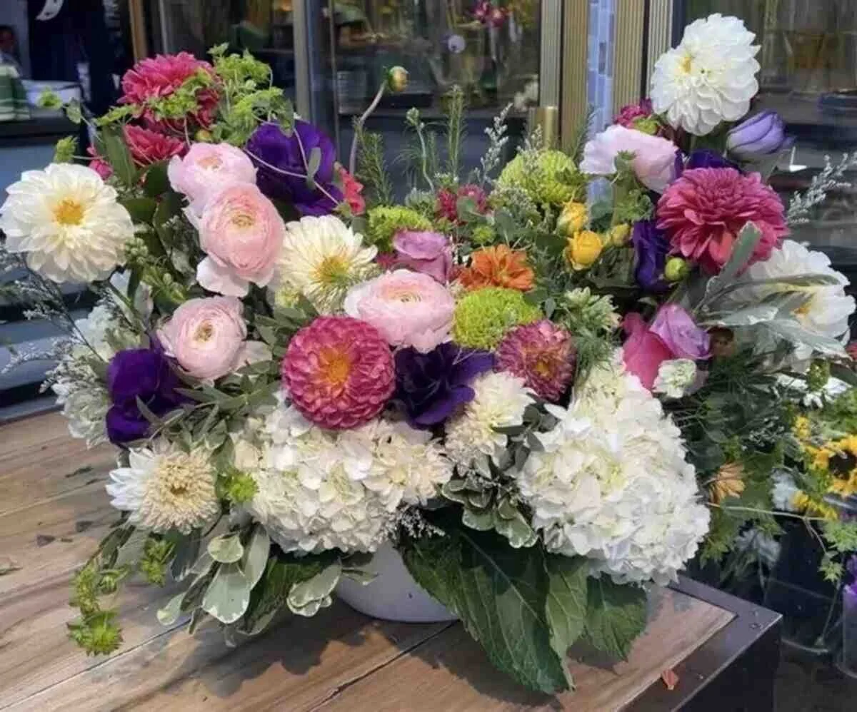 Colorful mixed flower arrangement with white hydrangeas, pink ranunculus, purple lisianthus, and other blooms in a white vase on a wooden table.