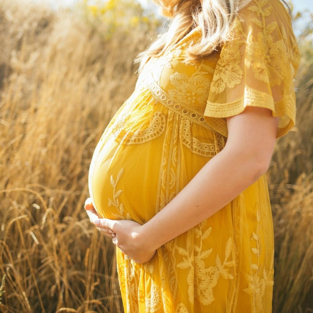 A pregnant woman in a yellow dress standing in a golden field, hands resting on her belly