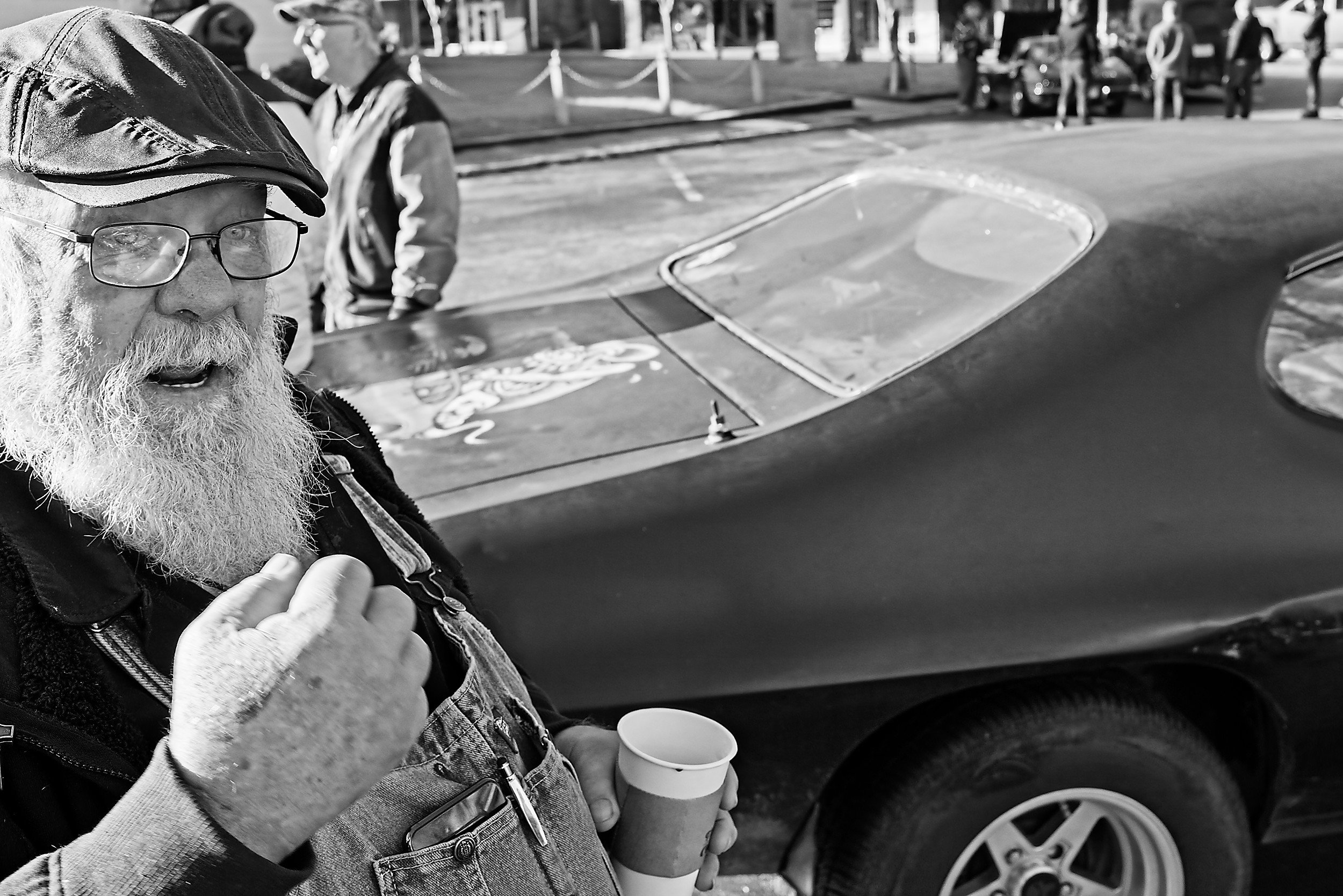  black and white image of a bearded man holding a paper coffee cup in front of a vintage car 