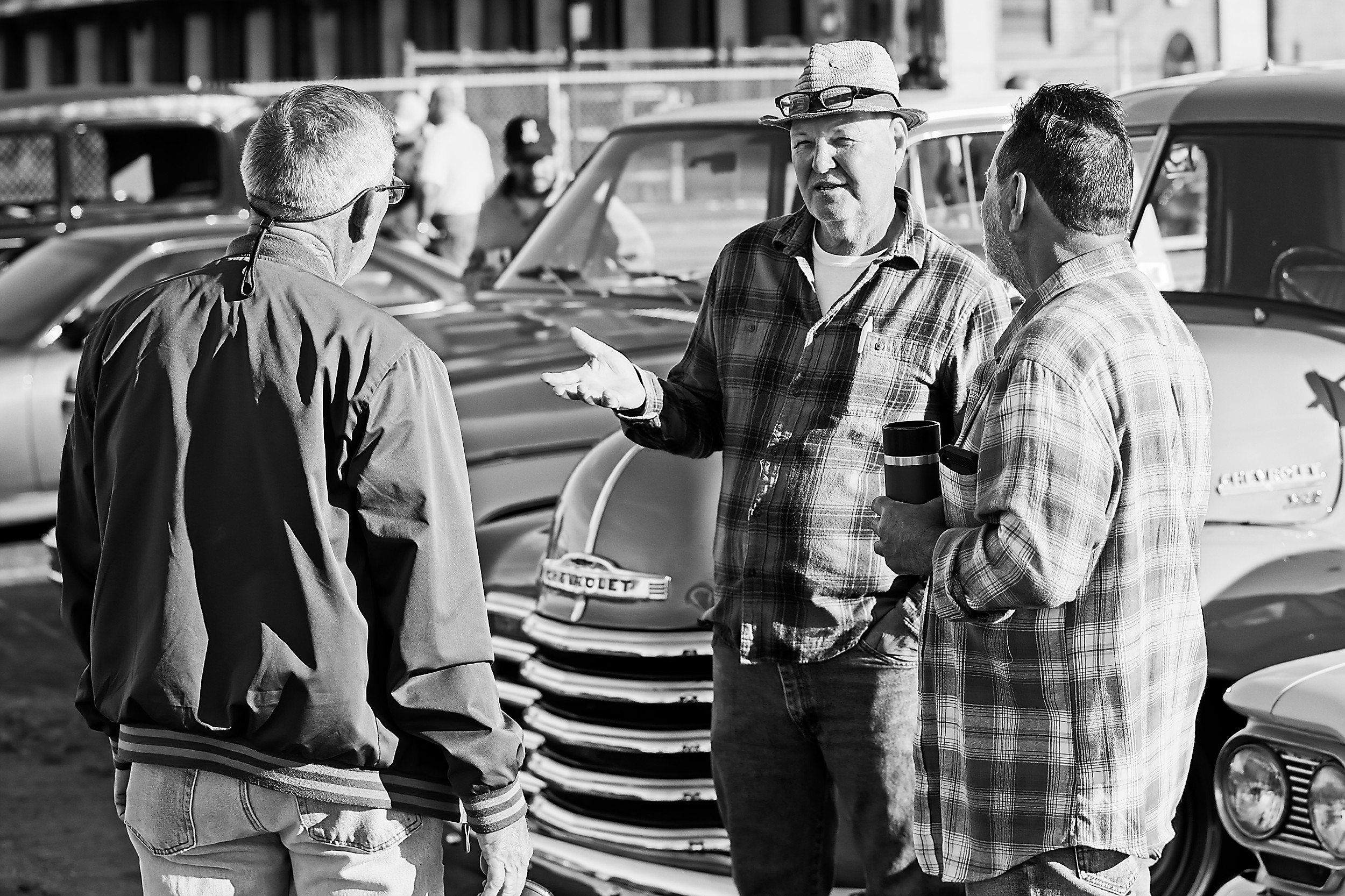  black and white image of three men talking in front of a vintage car at a car show 