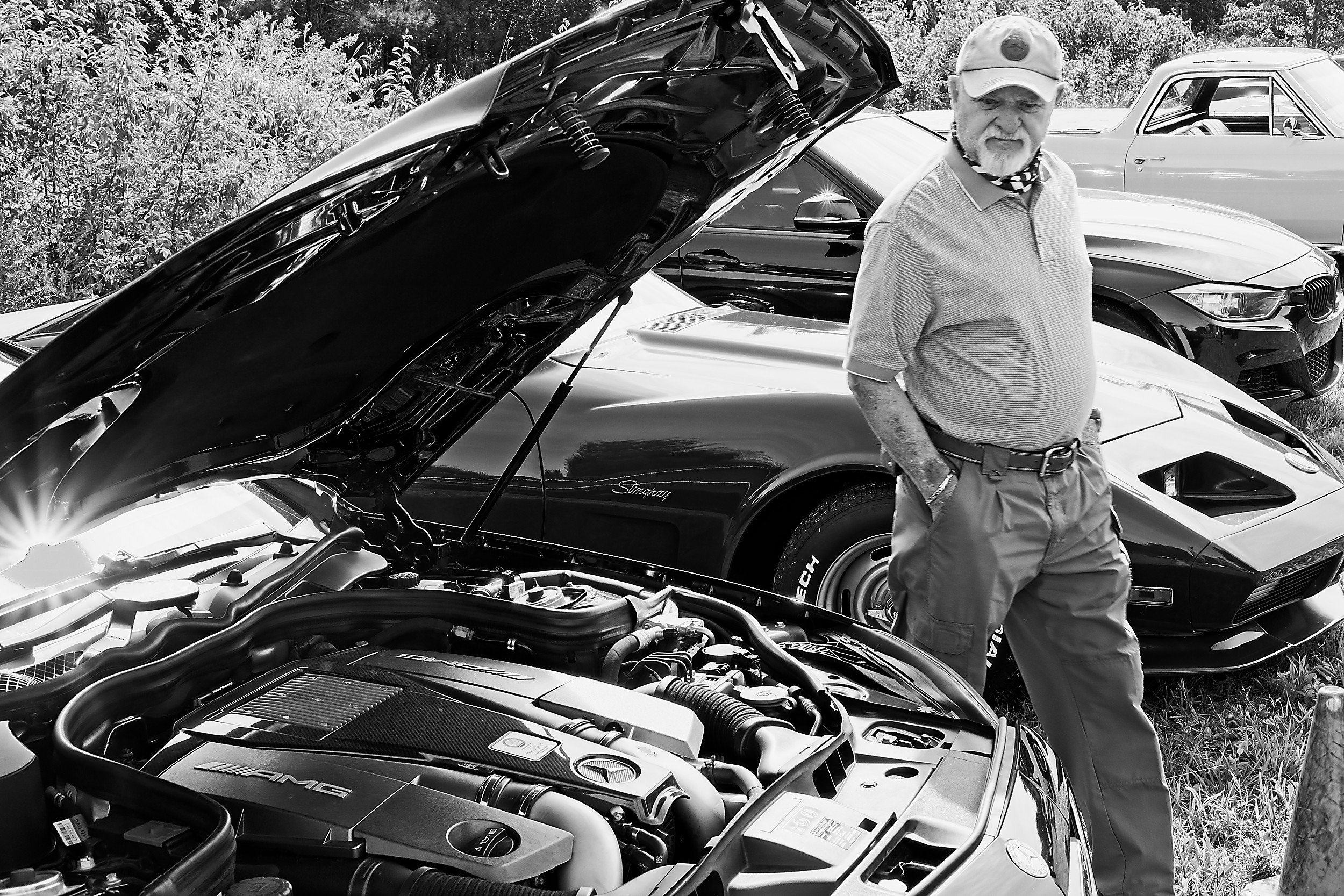  black and white image of a man passing by a vintage car and looking at the opened hood 