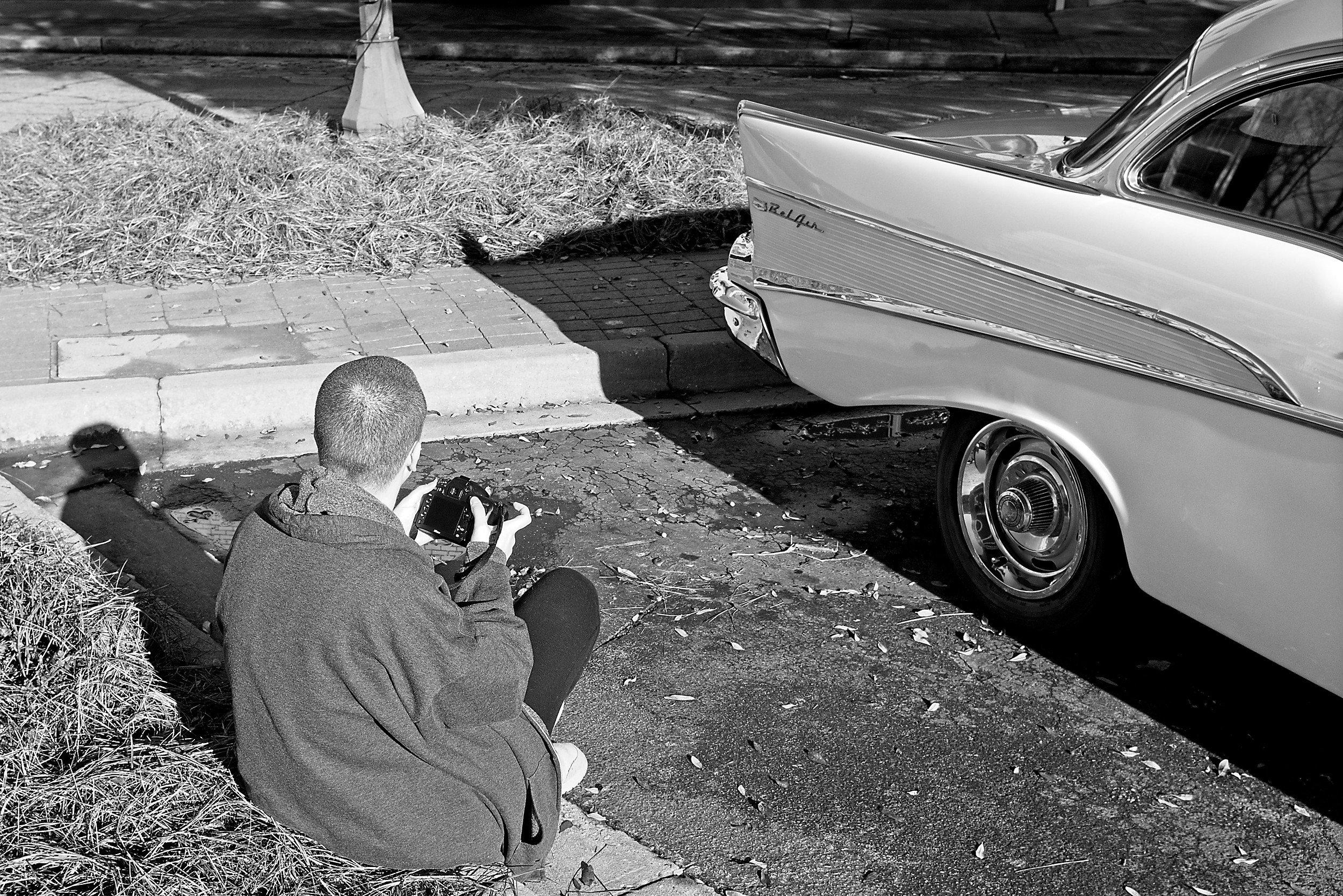  black and white image of a female photographer sitting on the sidewalk to photograph the details of a vintage car 