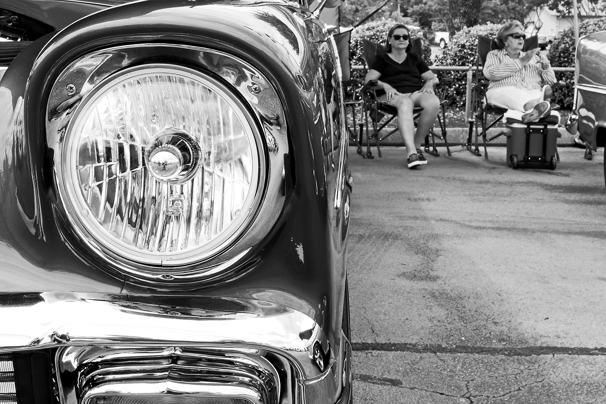  black and white close up image of the light of a vintage car with two women sitting in the background 