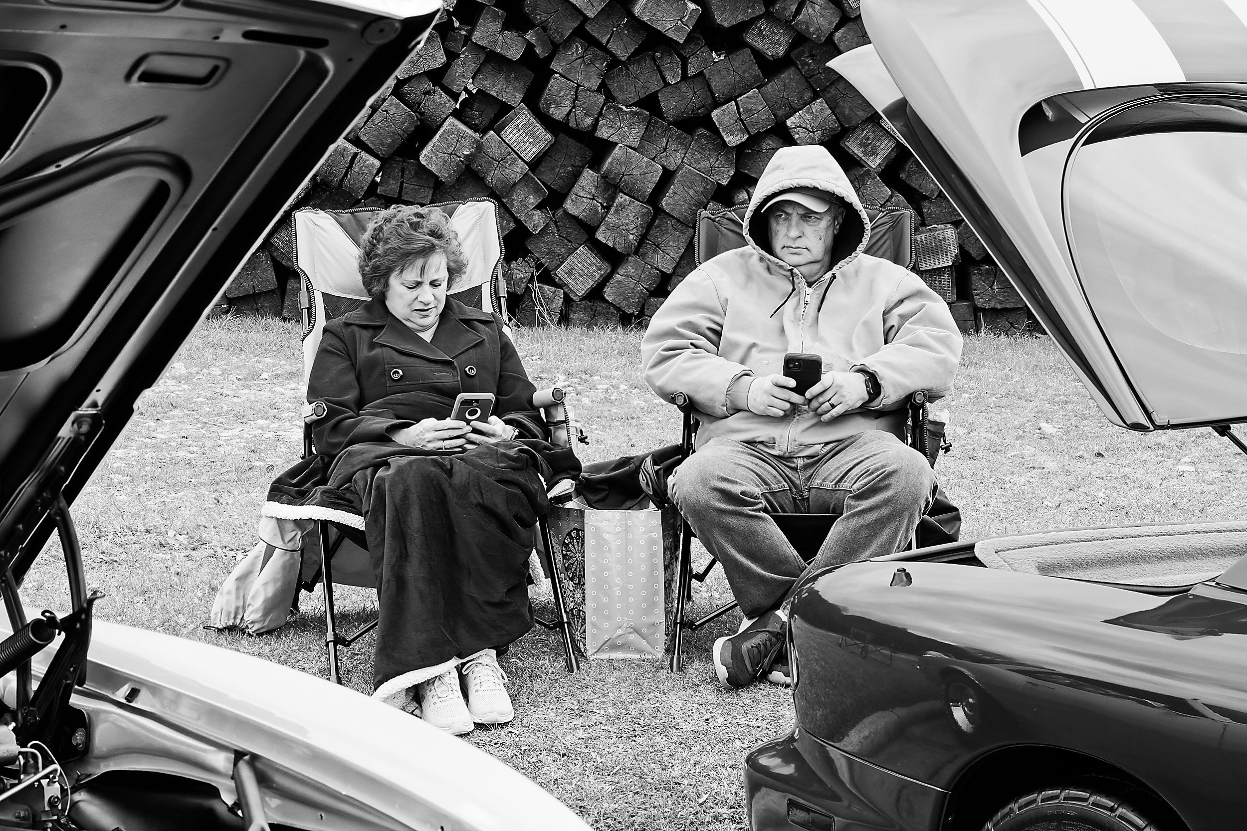  black and white image of a man and a woman sitting on foldable chairs at a vintage car show 