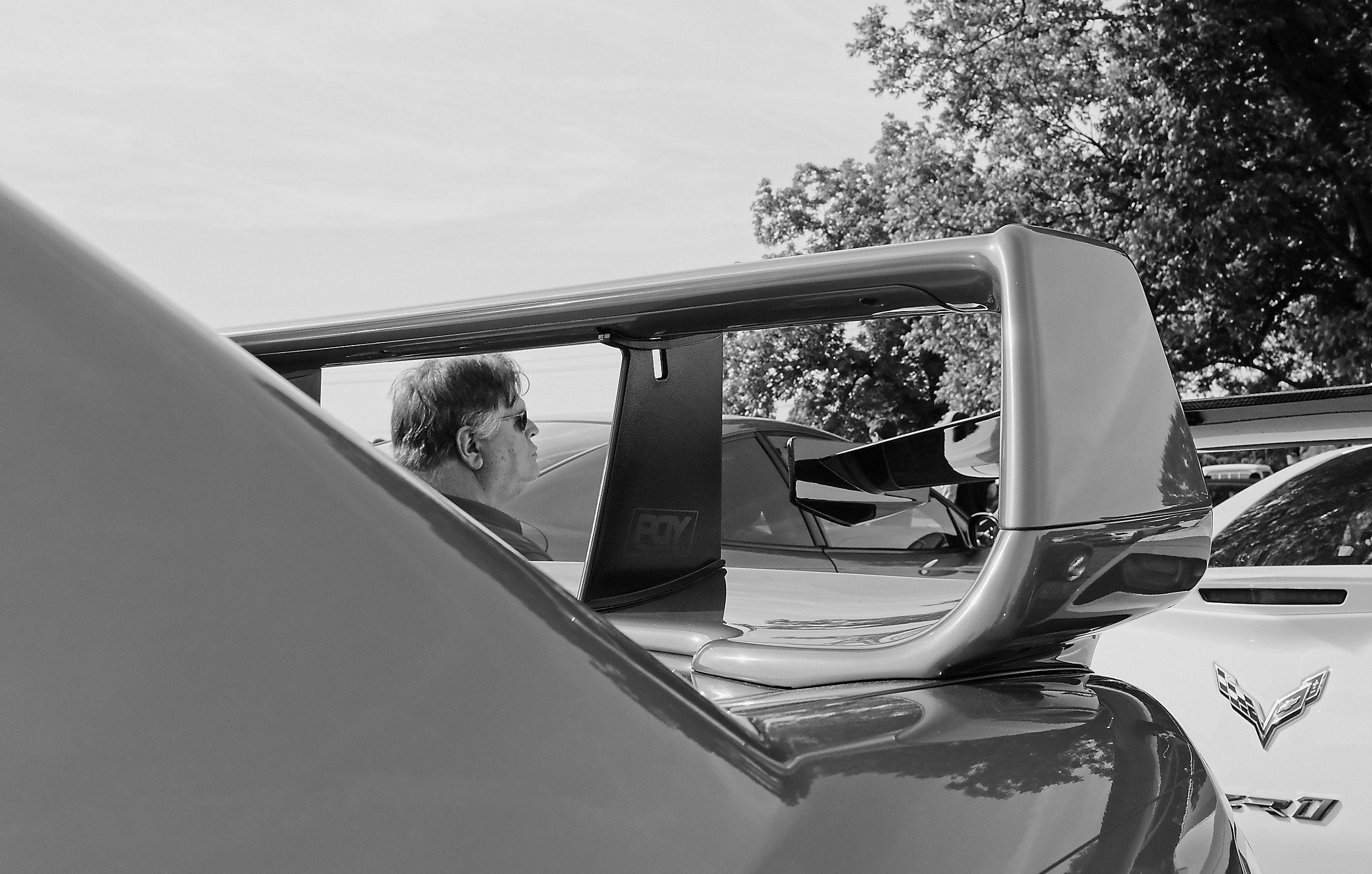  black and white image of the head of a man as seen through the wing of a car at a car show 