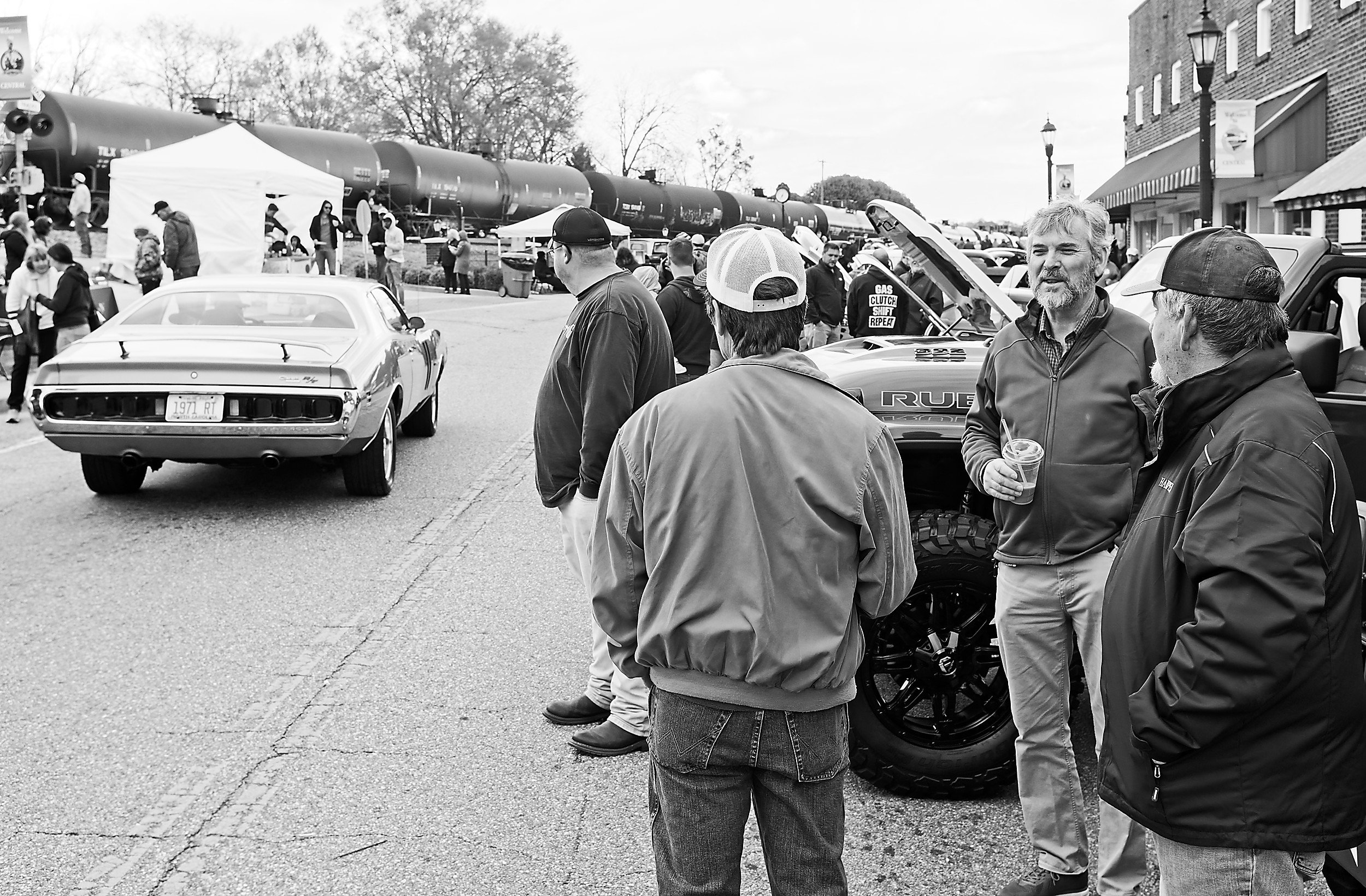  black and white image of several men looking at a car driving away at a car show 