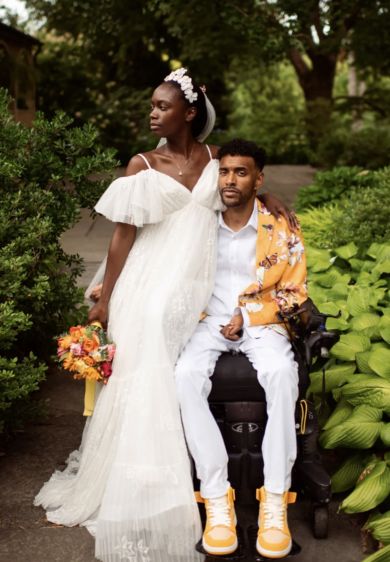 A black woman in a white wedding dress stands close to a black man in a suit and yellow trainers, who is in a wheelchair.