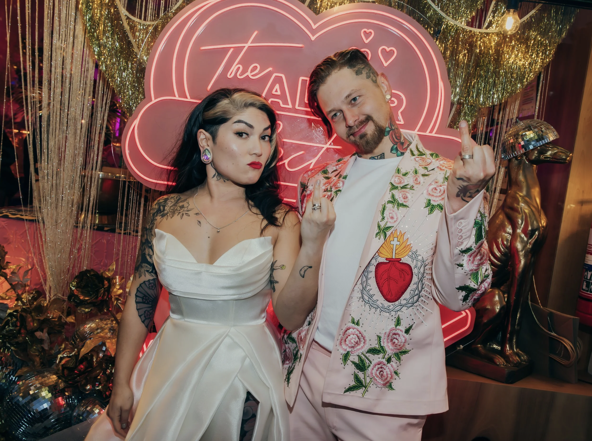 A bride and groom stand in front of a neon altar sign and both show off their ring fingers with new wedding rings.