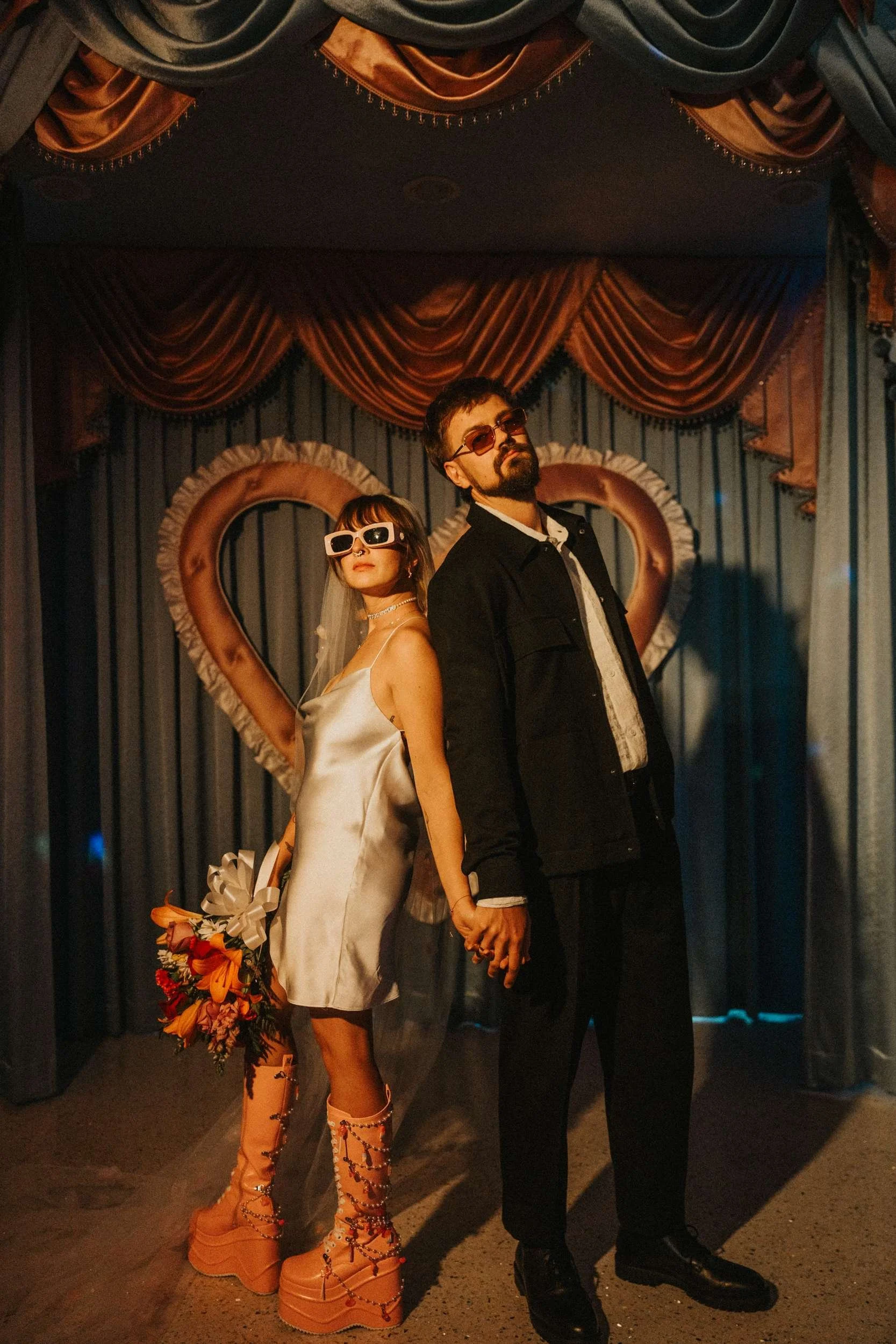 A bride in a short white satin dress and a groom in a casual suit both wear sunglasses and stand in front of baby blue curtains and a big pink heart.