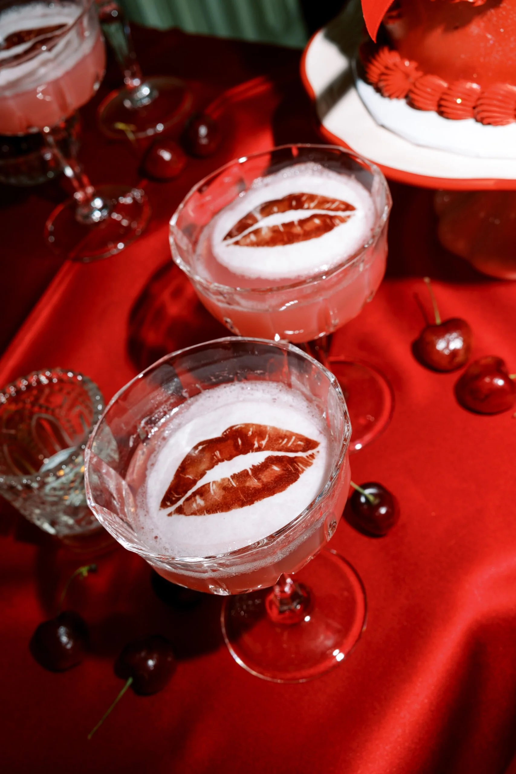Glasses of pink liquid with lipstick-kiss toppers lay on a red tablecloth, surrounded by cherries.