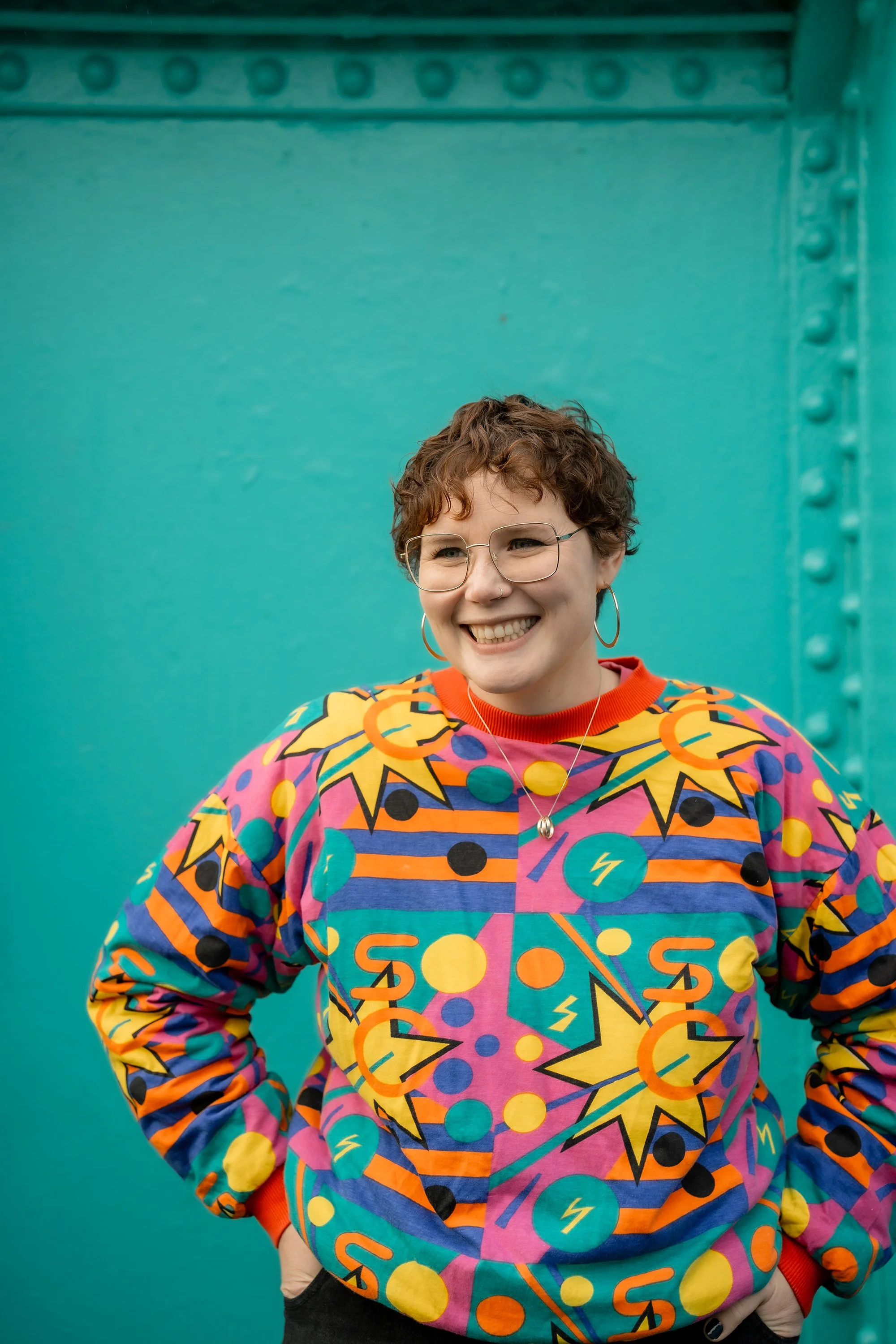 A white woman smiles just away from the camera. She wears gold hoop earrings, gold-rimmed glasses and a bright patterned jumper. She stands in front of a turquoise background.