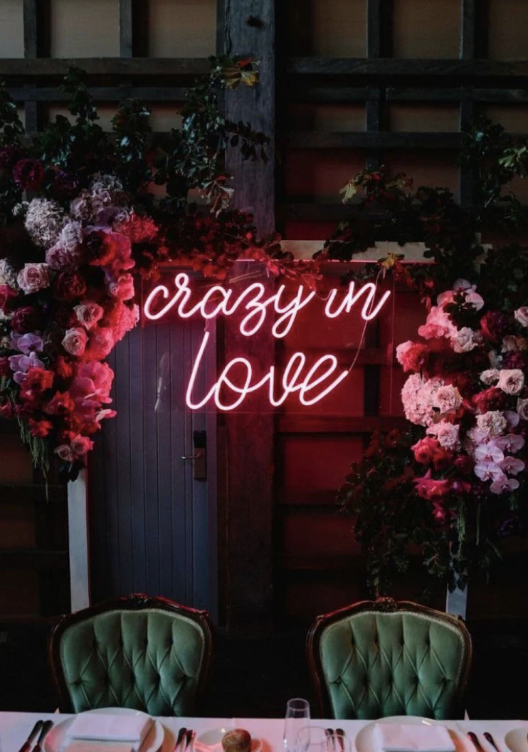 Neon pink sign which says 'crazy in love' behind a table, chairs and surrounded by flowers.
