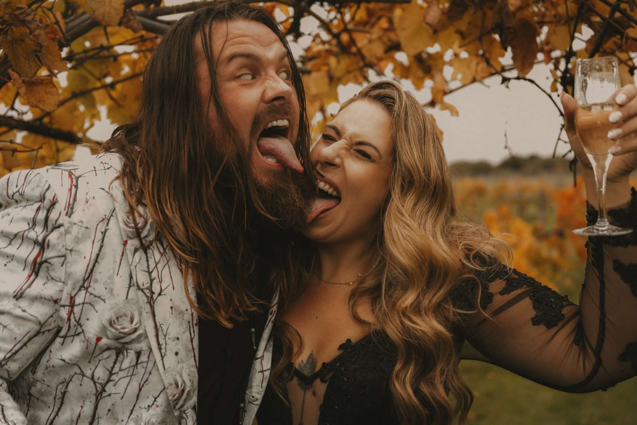 A man with a beard and long hair and a woman in a black dress stick their tongues out at one another.