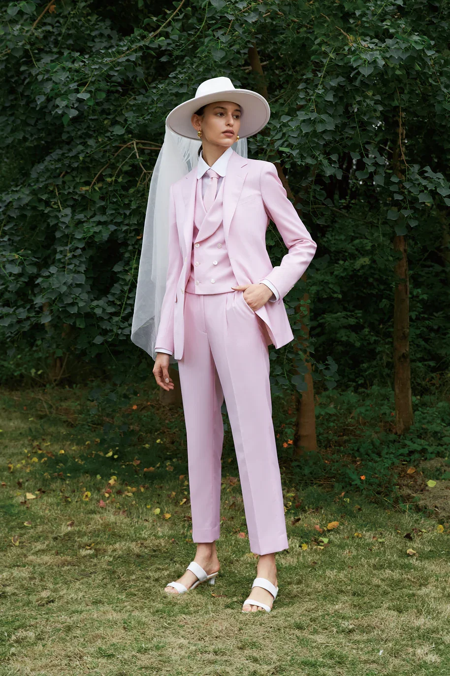 A person in a pink three-piece suit, fedora hat and veil stands on grass in front of trees.