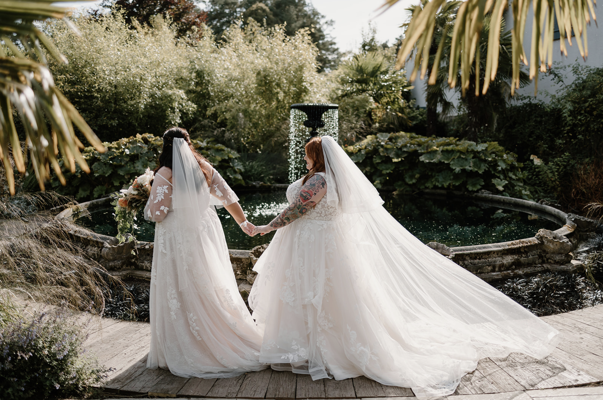 Two brides walk hand-in-hand around a beautiful waterfall in an English garden.