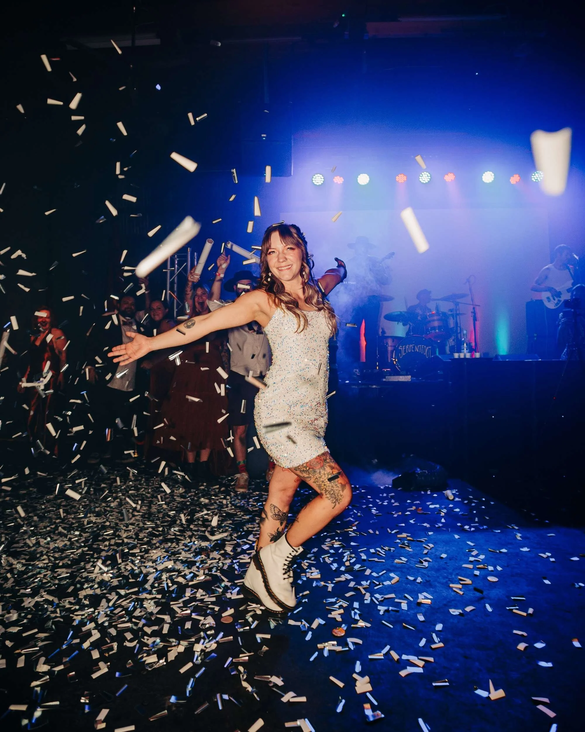 A bride in a short white sequin dress and white Doc Martens holds her arms out on a confetti-filled dance floor with a live band on a stage behind her.