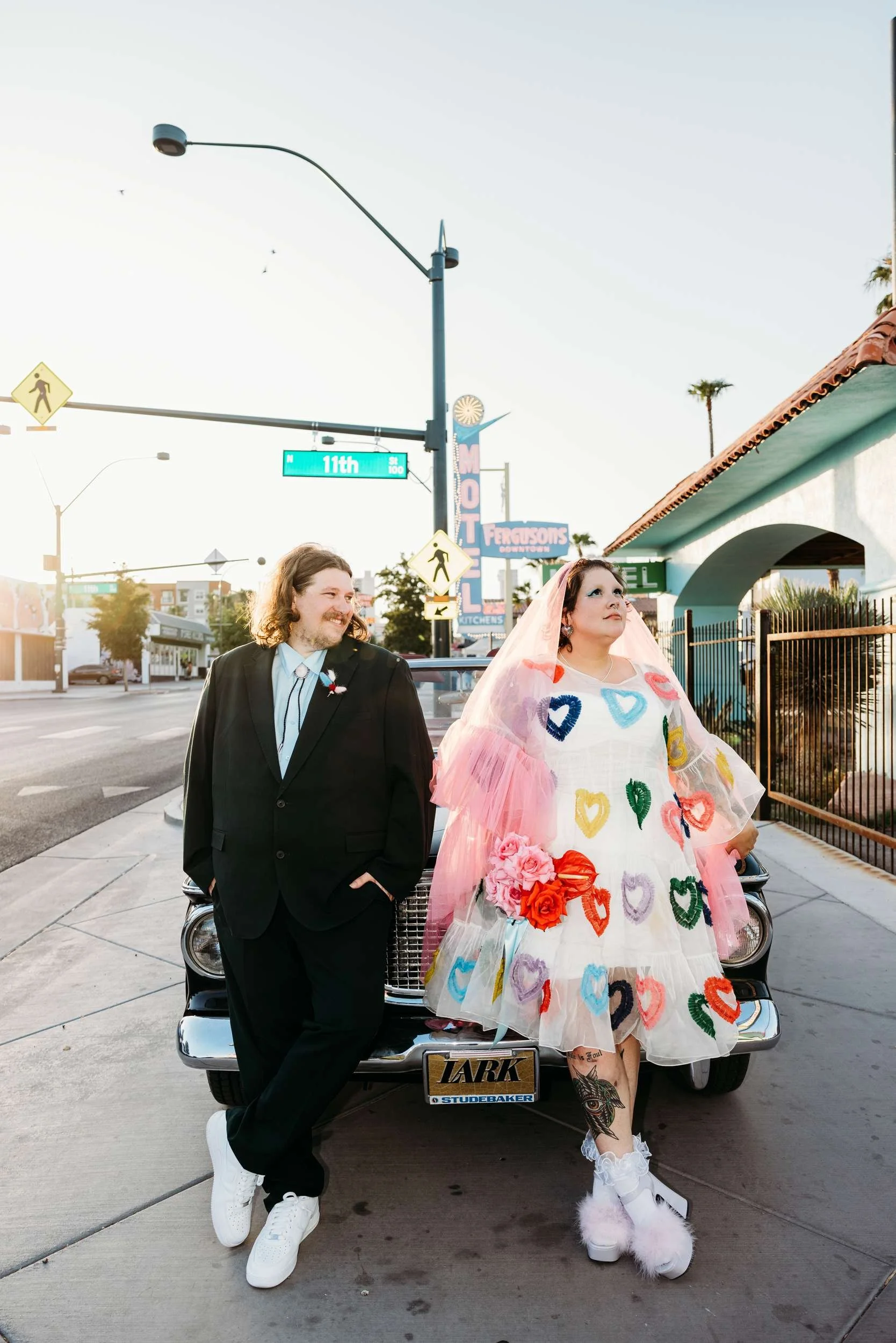 Two big-bodied folk look stunning and happy posed against a vintage car. The bride wears a white dress covered in colourful hearts and a pink tulle veil.