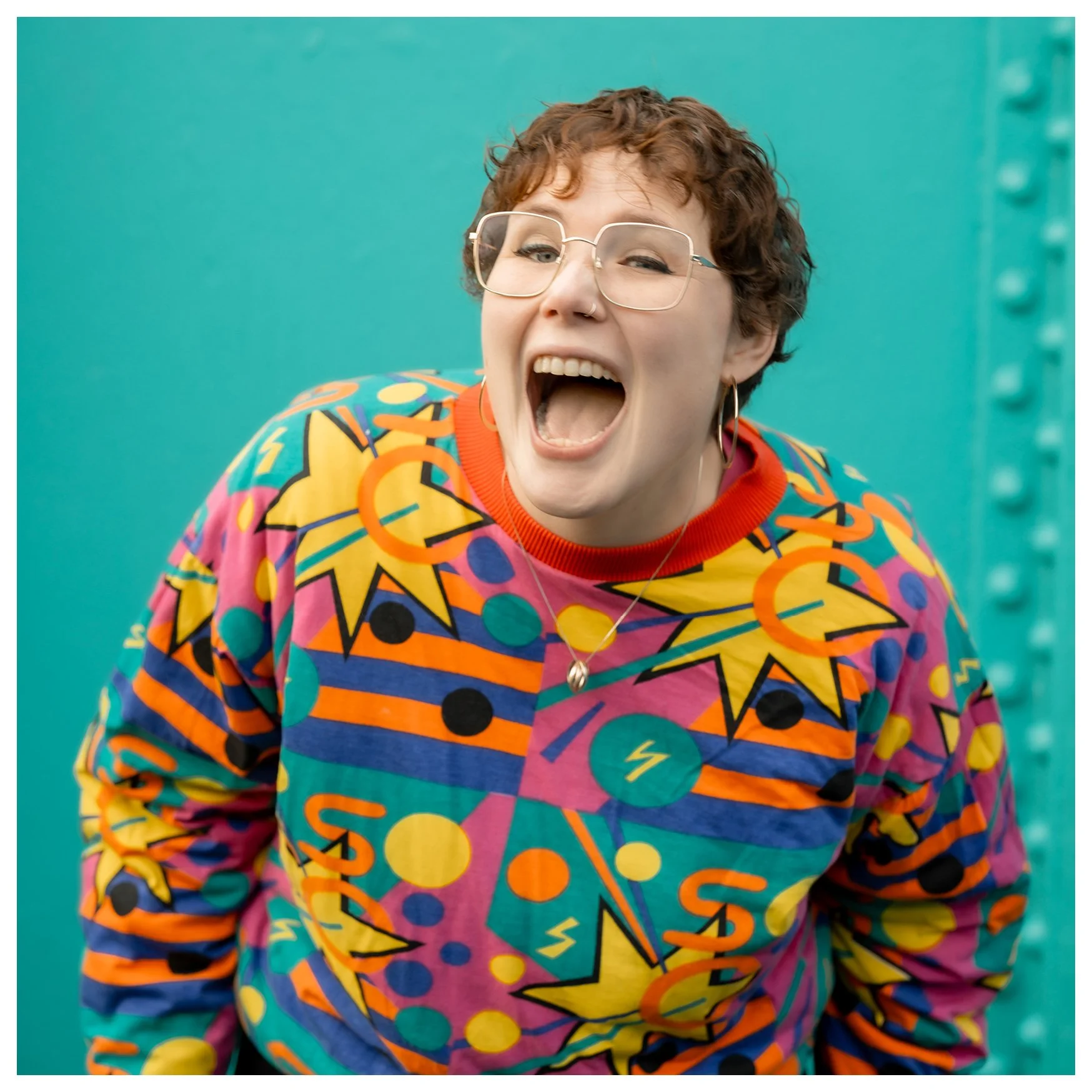 A white woman laugh directly at the camera. She wears gold hoop earrings, gold-rimmed glasses and a bright patterned jumper. She stands in front of a turquoise background.