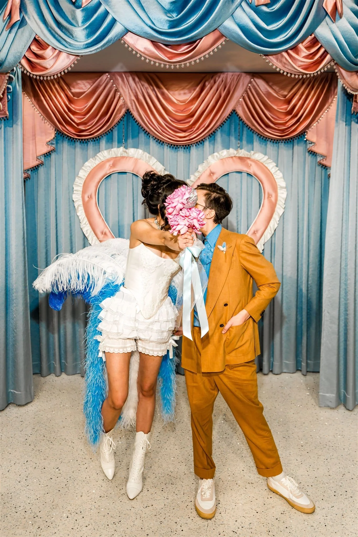 A bride and groom kiss behind a bouquet whilst standing in front of a satin draped background with a big heart on the wall.