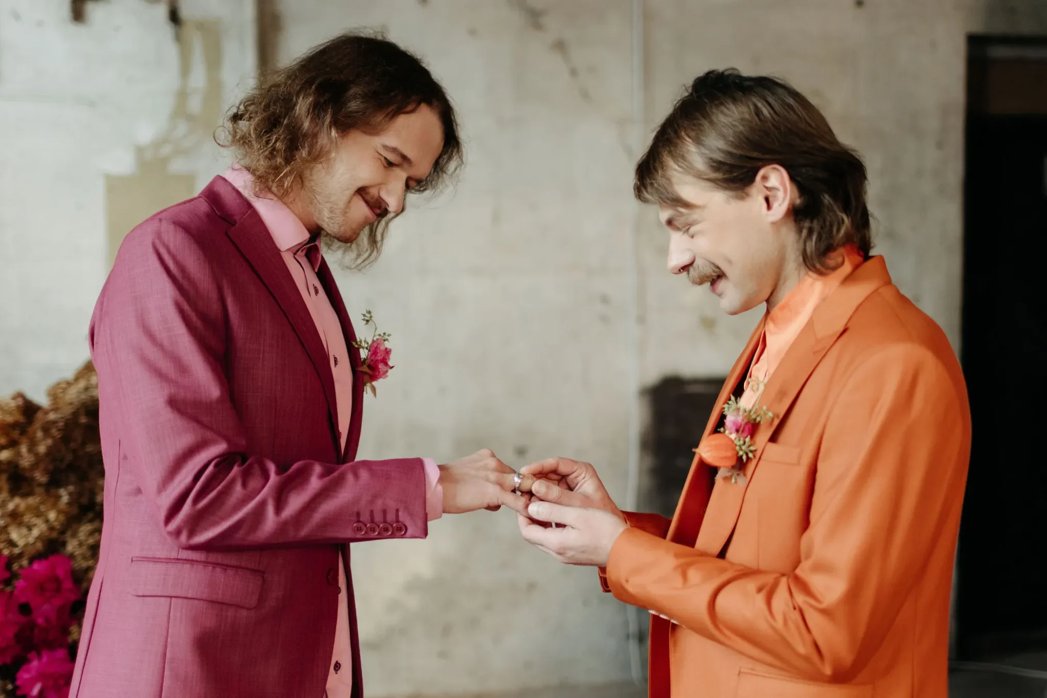 Two white males with moustaches, one in an orange suit and the other in cranberry pink, exchange rings in front of a flower arch.