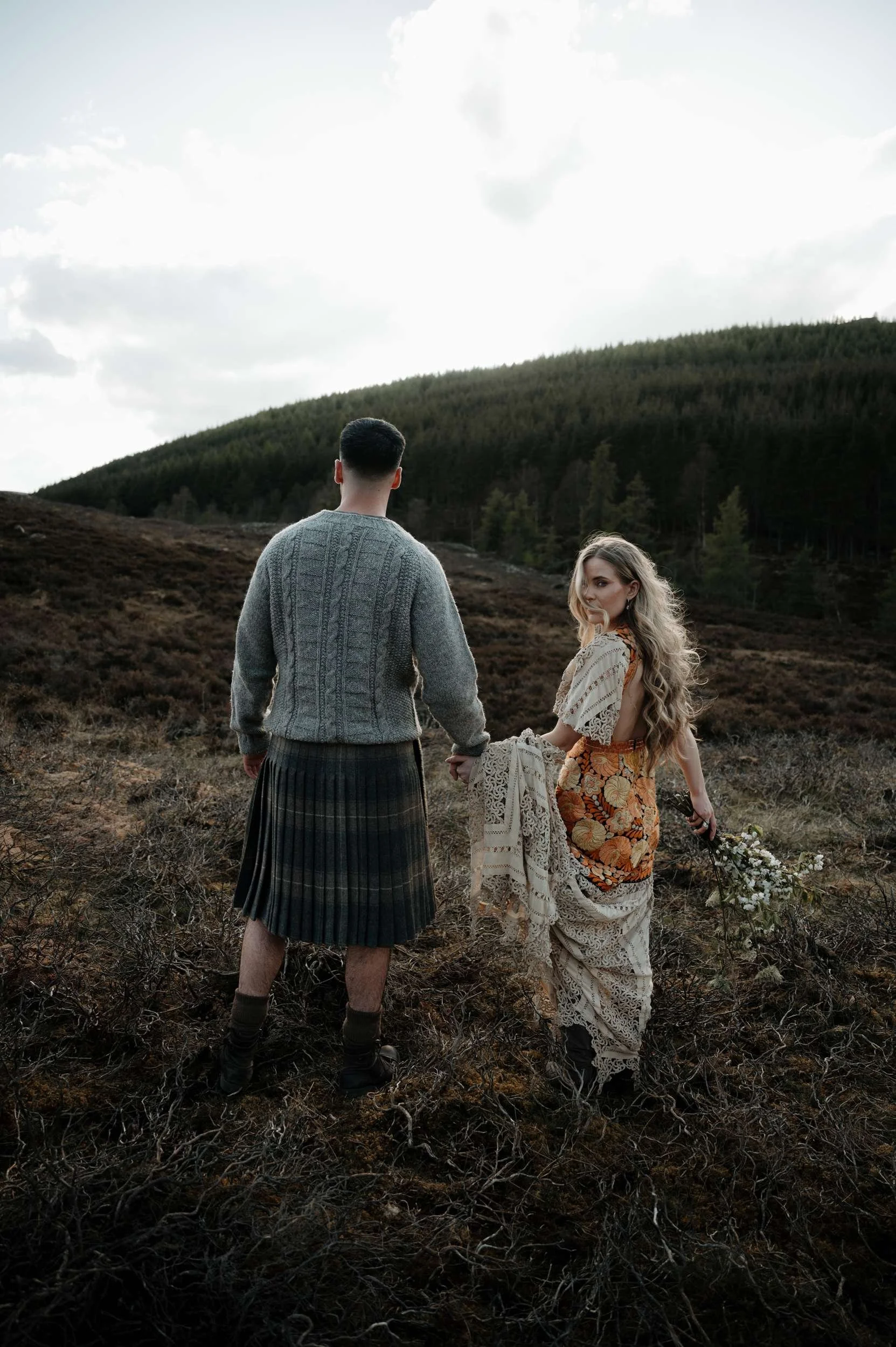 A man and woman hold hands in a mountain scene. The man has his back to us but the woman, who wears a boho-style dress, looks over her right shoulder at the camera.