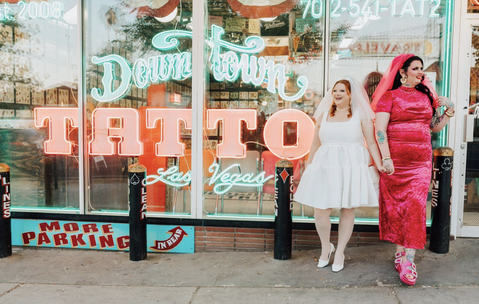 Two brides, one in white and one in hot pink hold hands in front of the neon sign of a Las Vegas tattoo studio.