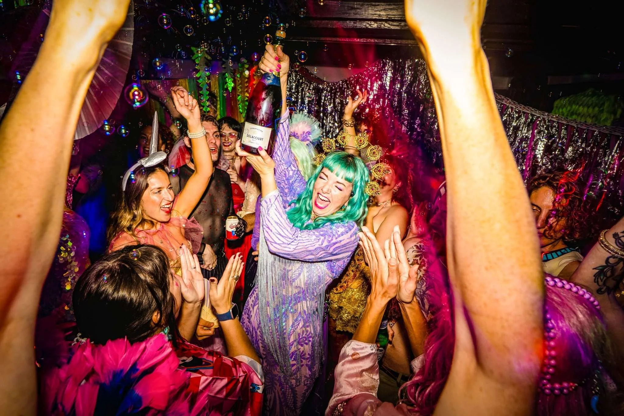 A woman in a sequin purple outfit with green hair holds up a bottle of champagne surrounded by cheering friends.