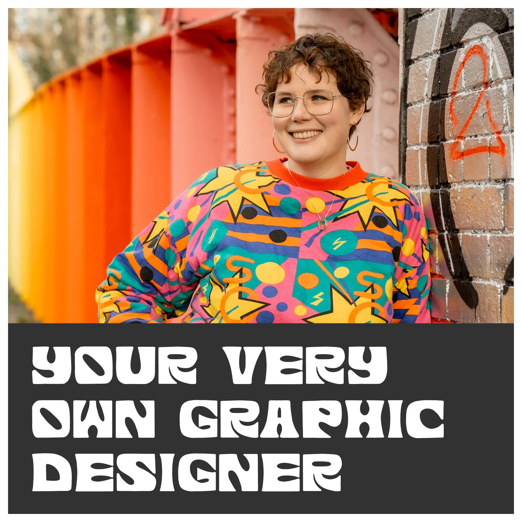 A white woman smiles just away from the camera. She wears gold hoop earrings, gold-rimmed glasses and a bright patterned jumper. She stands in front of a rainbow bridge and a graffitied wall.