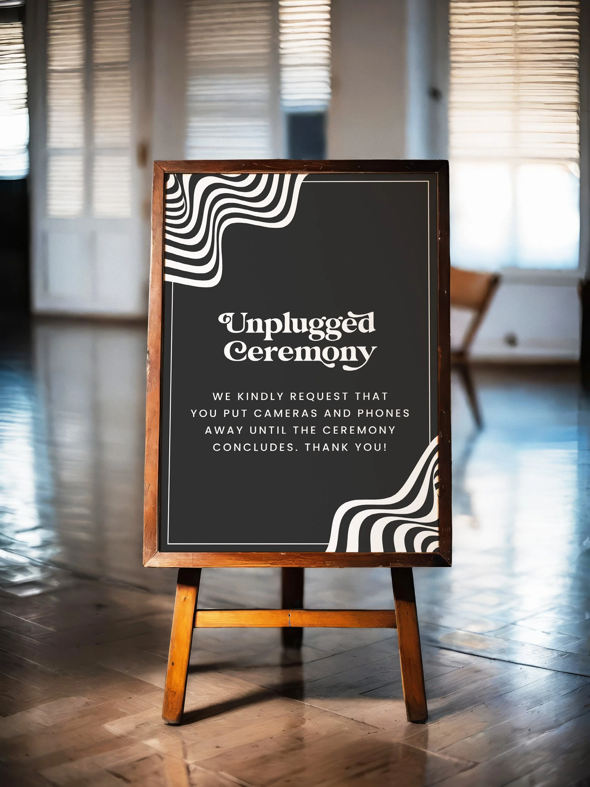A large easel and sign in the middle of an inductrial wedding venue. The sign is black with white retro patterns and asks guests to keep phones and cameras away until the end of the ceremony.