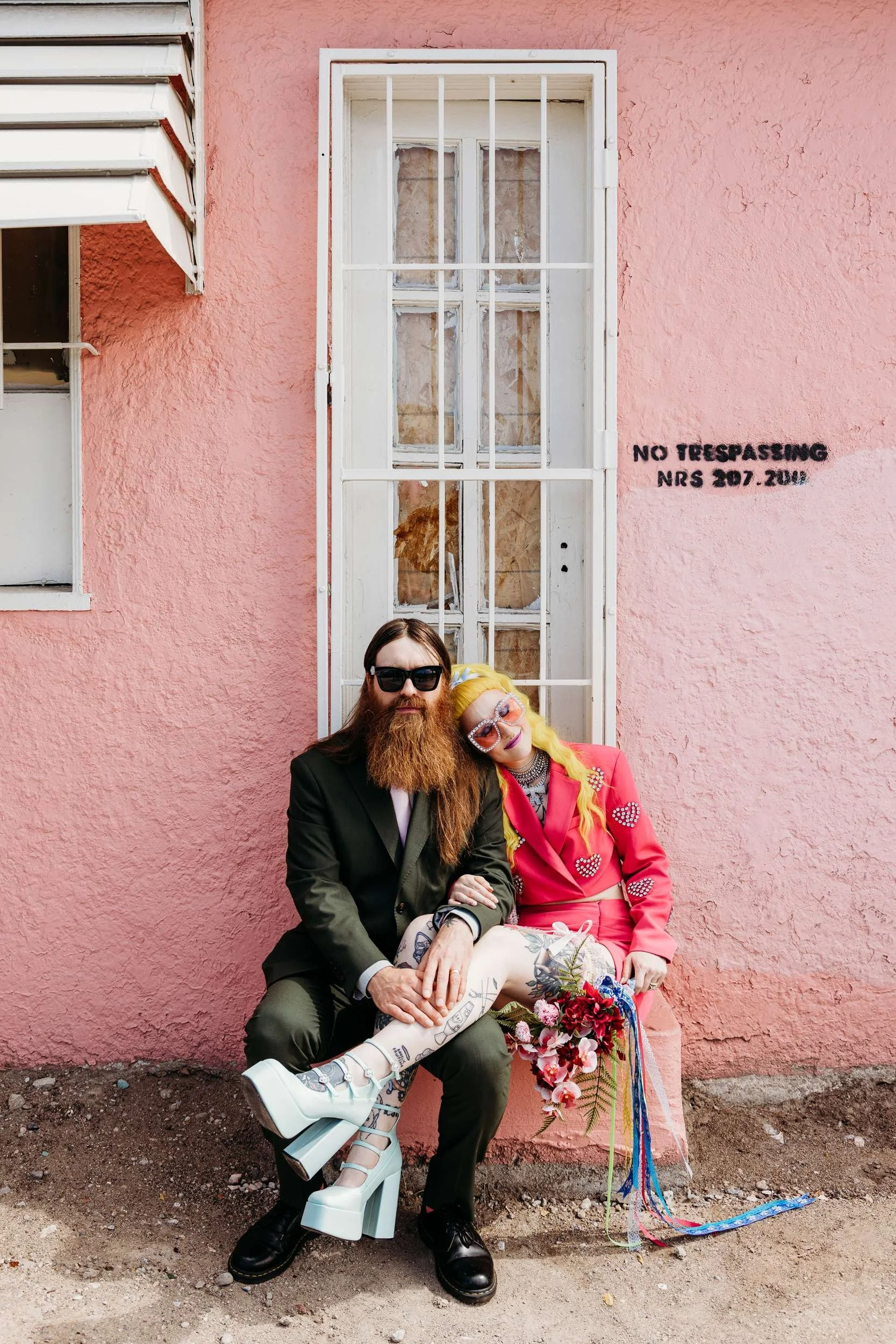 A woman in a pink bejewelled two piece and diamond glasses leans her head towards a beared man in a suit and Doc Martens in front of a pink wall.