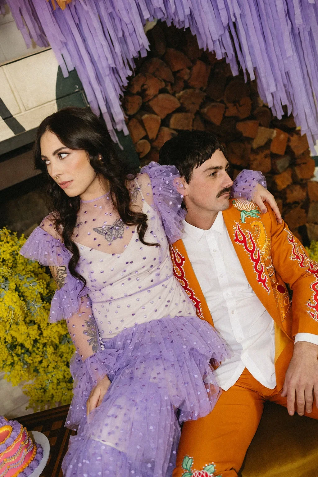A bride with a nose piercing and polkadot purple dress sits with her new husband, who wears an orange suit embroidered with flames and flowers.