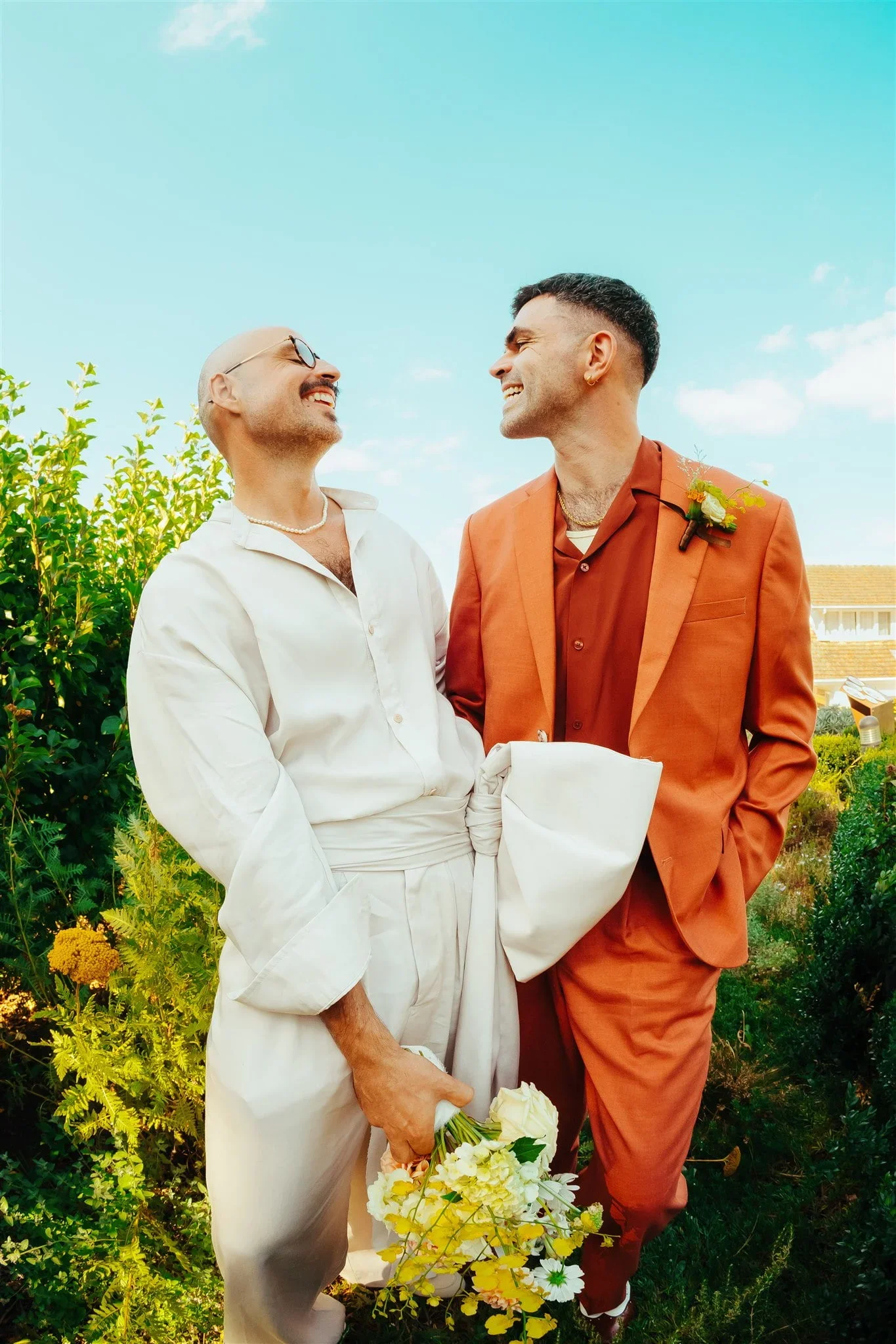 A pair of lovers smaile widely at one another in an orange suit and a white two-piece, and the photo is taken with a wide-angle lens.