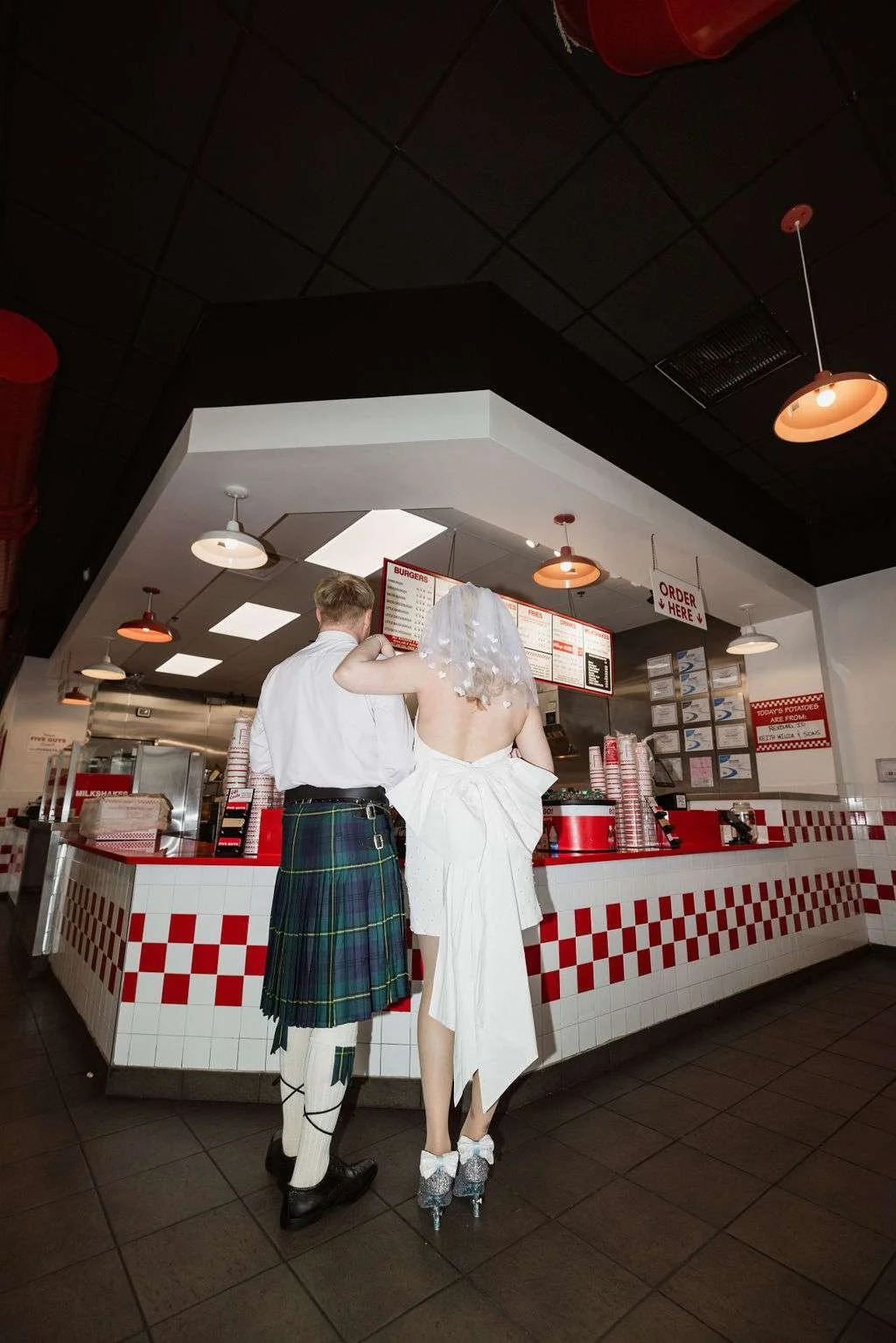 We see a man in a kilt and a woman in a short white dress and veil ordering burgers in a Five Guys.