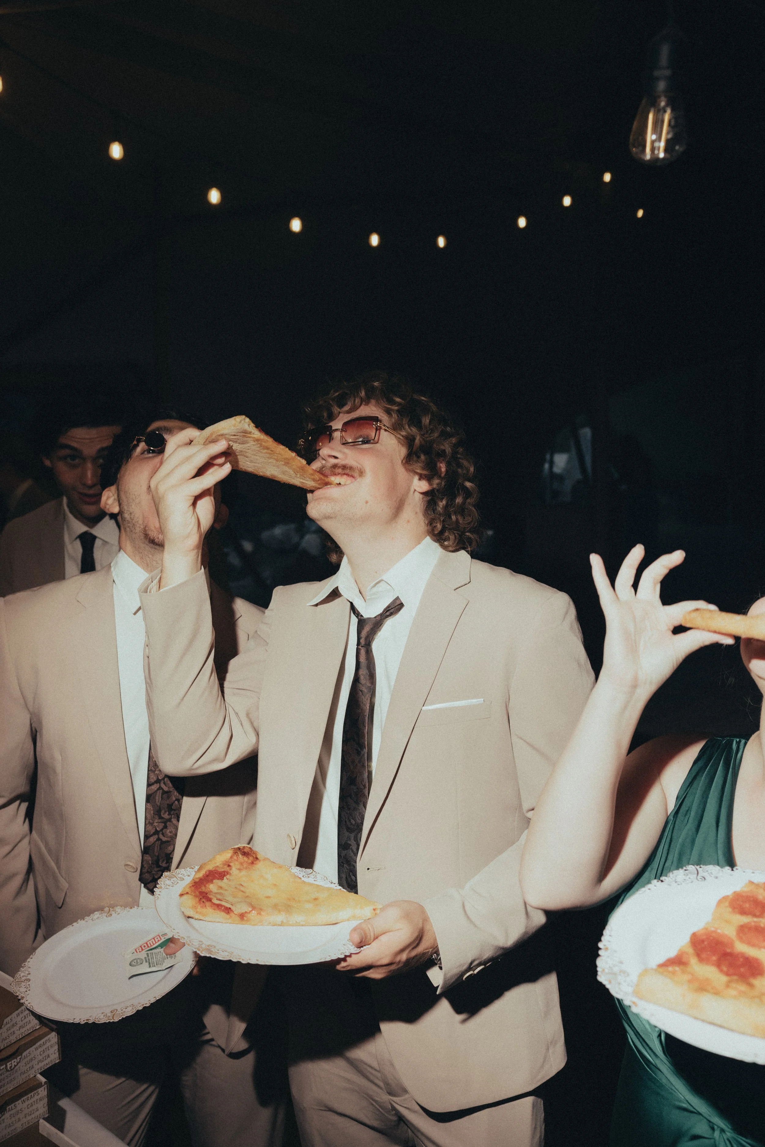 A man in a light suit smiles at the camera whilst eating pizza.
