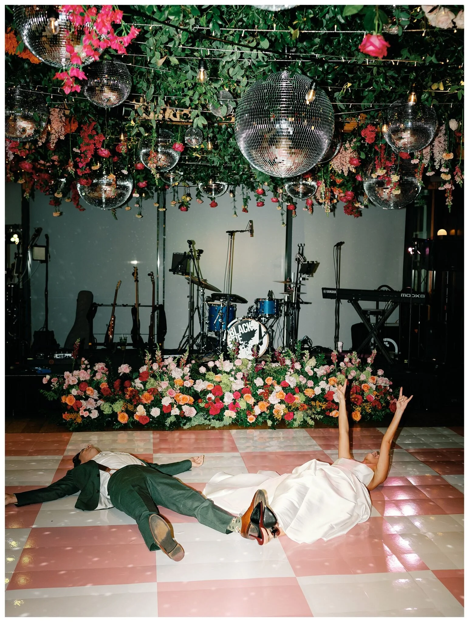 A man and woman lay on a pink and white chequered dance floor in front of a drum kit.