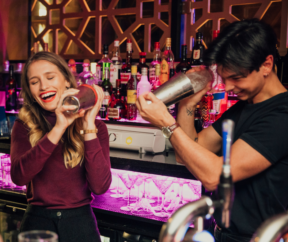 A white woman and mixed race man shake cocktails behind a bar whilst laughing.