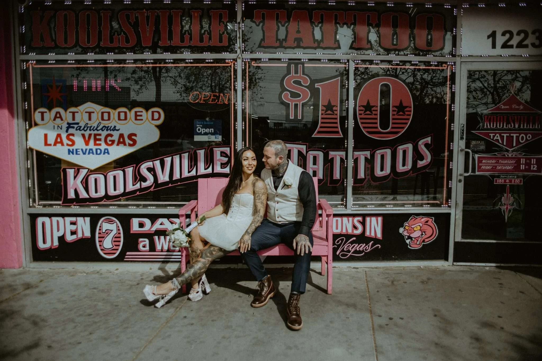 A woman in a white dress and platforms with tattoos on her arms and legs leans against a man also covered in tattoos on a pink bench outside a tattoo parlour.