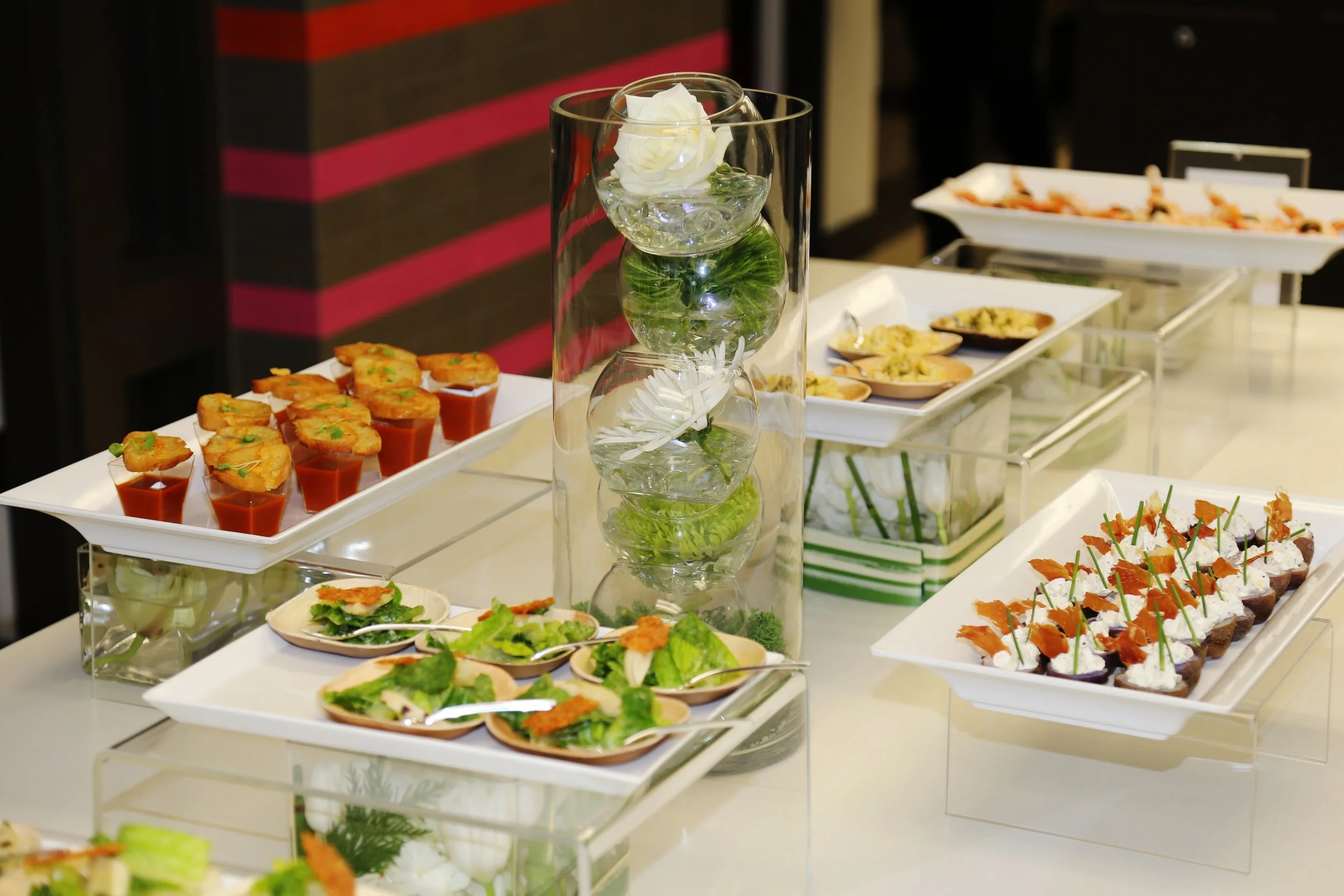Elegant buffet table with various appetizers and desserts including canapés, salads, and small dishes, along with a glass hurricane candle holder with white flowers inside.